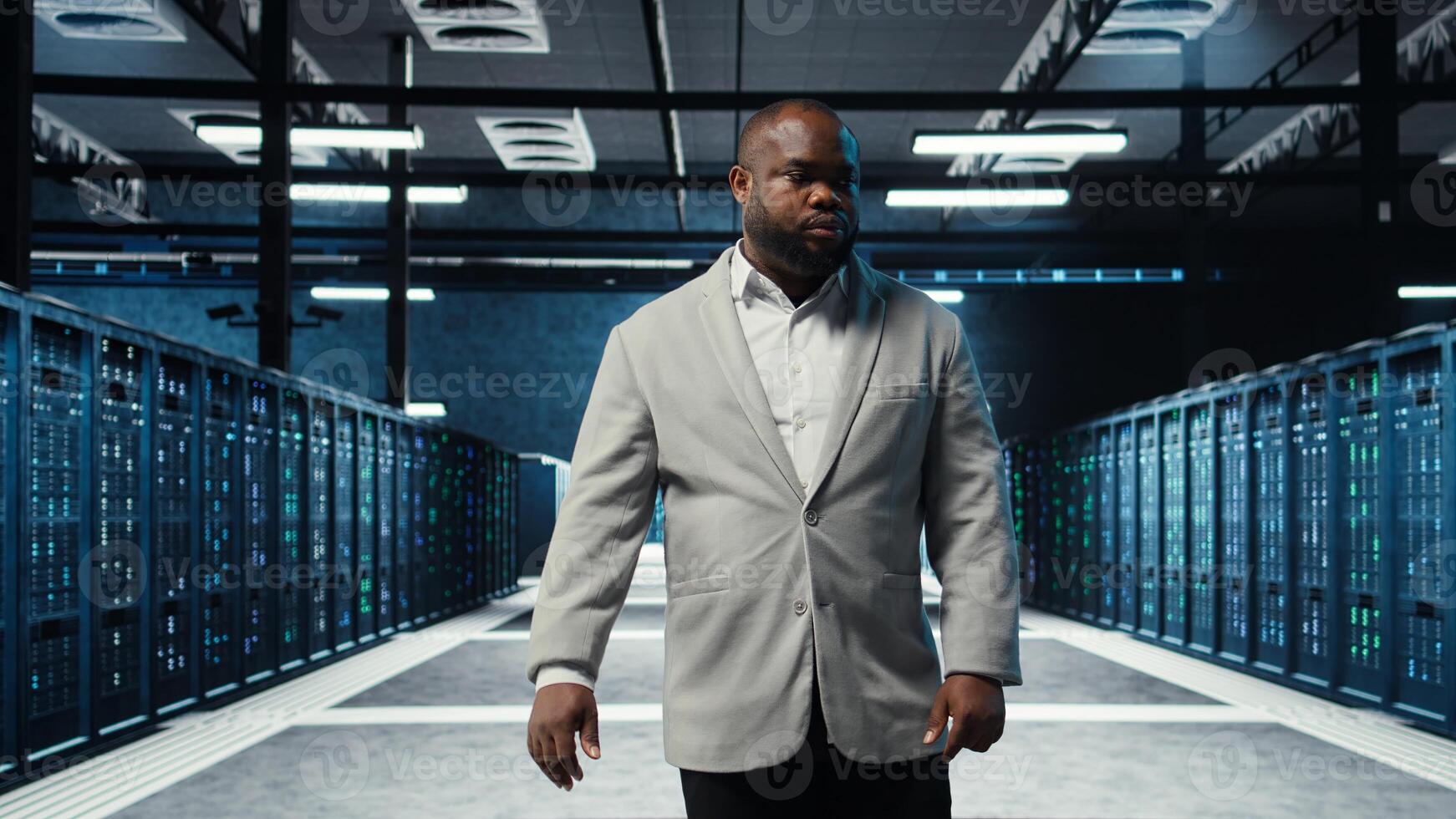 Employee walking in data center with blinking lights on rigs showing system activity. IT professional overseeing dimly lit server farm with equipment arranged for smooth operations, camera A photo