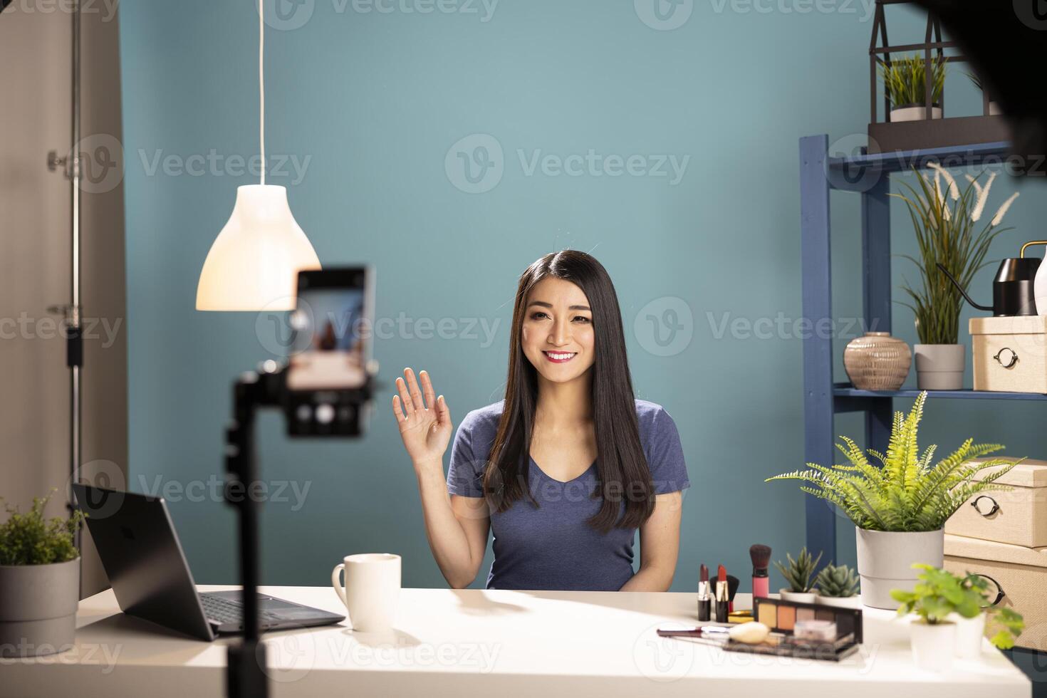 Beauty influencer waves at a mobile device, preparing to review cosmetics at her studio desk. Asian female vlogger records a makeup tutorial for her online audience using a tripod mounted smartphone photo