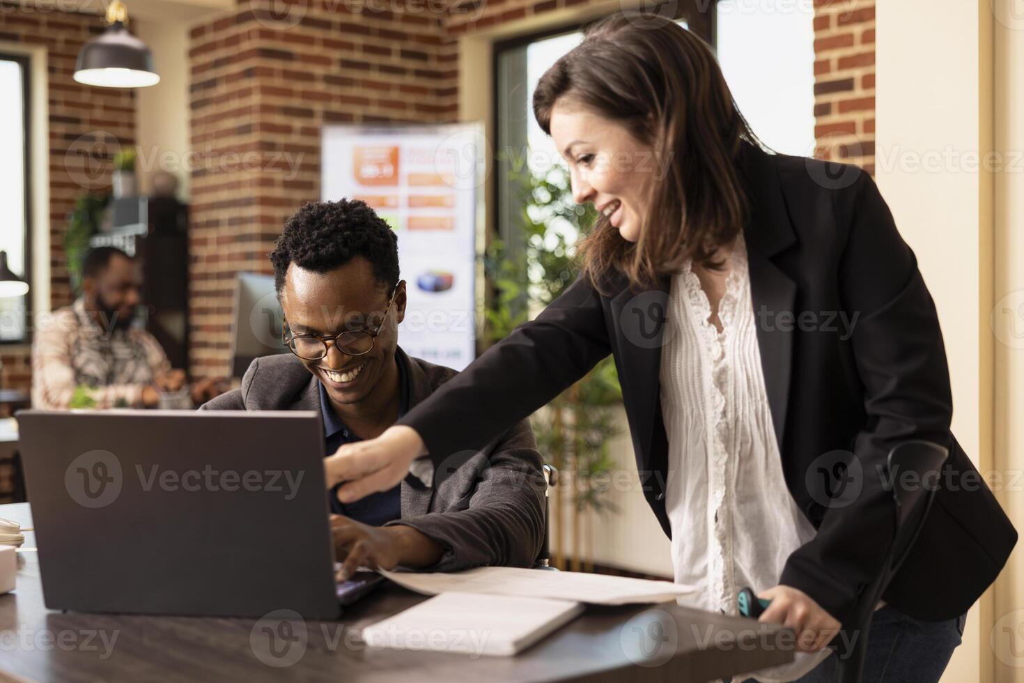 Black man sharing a laughter with his caucasian female coworker at the office desk while having a business discussion. Cheerful multiethnic colleagues having a meeting, reviewing reports on laptop. photo