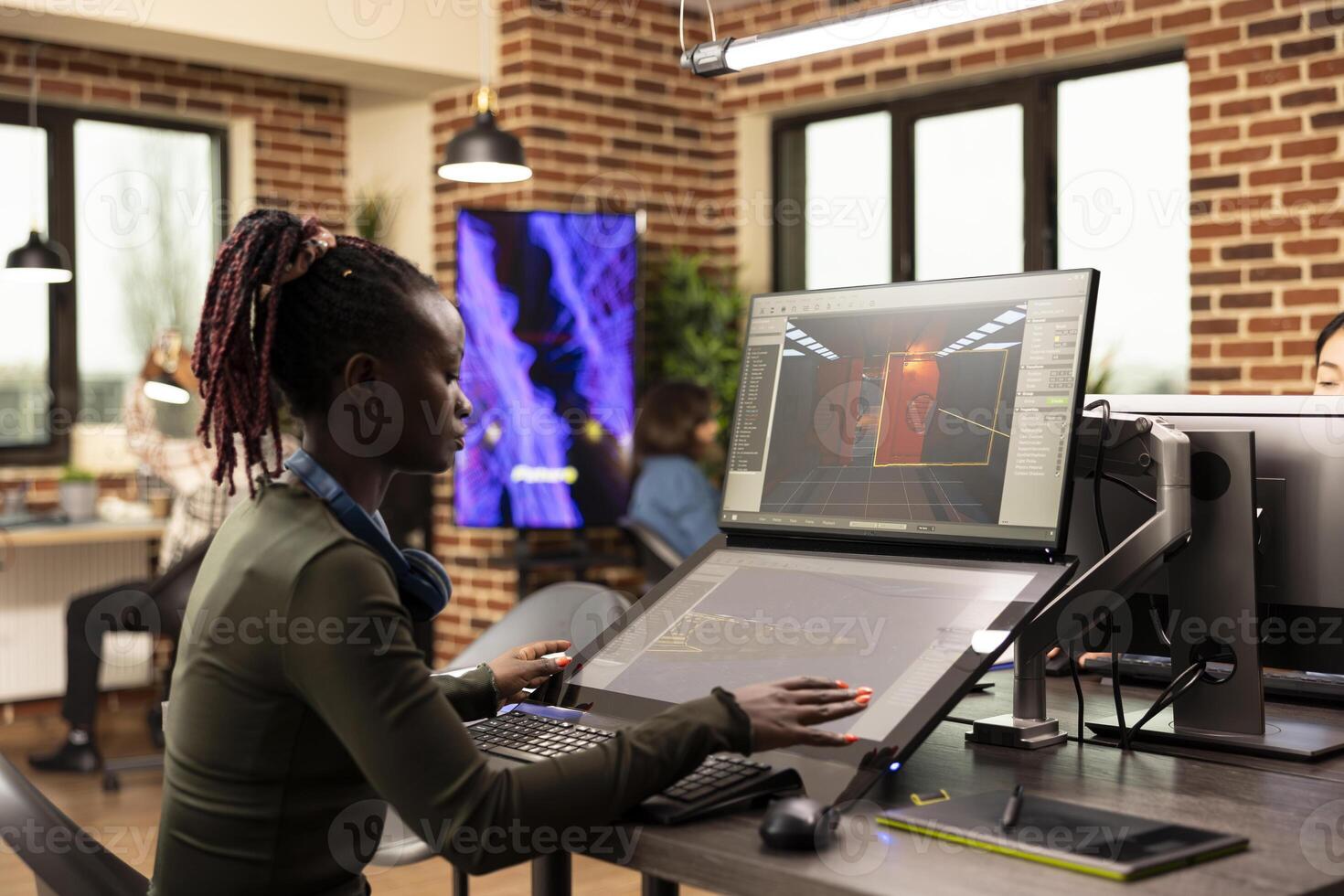 Focused female designer working on refining the graphics on her project in brick wall office. African american woman with headphones, analyzing product designs on multiple digital screens. photo