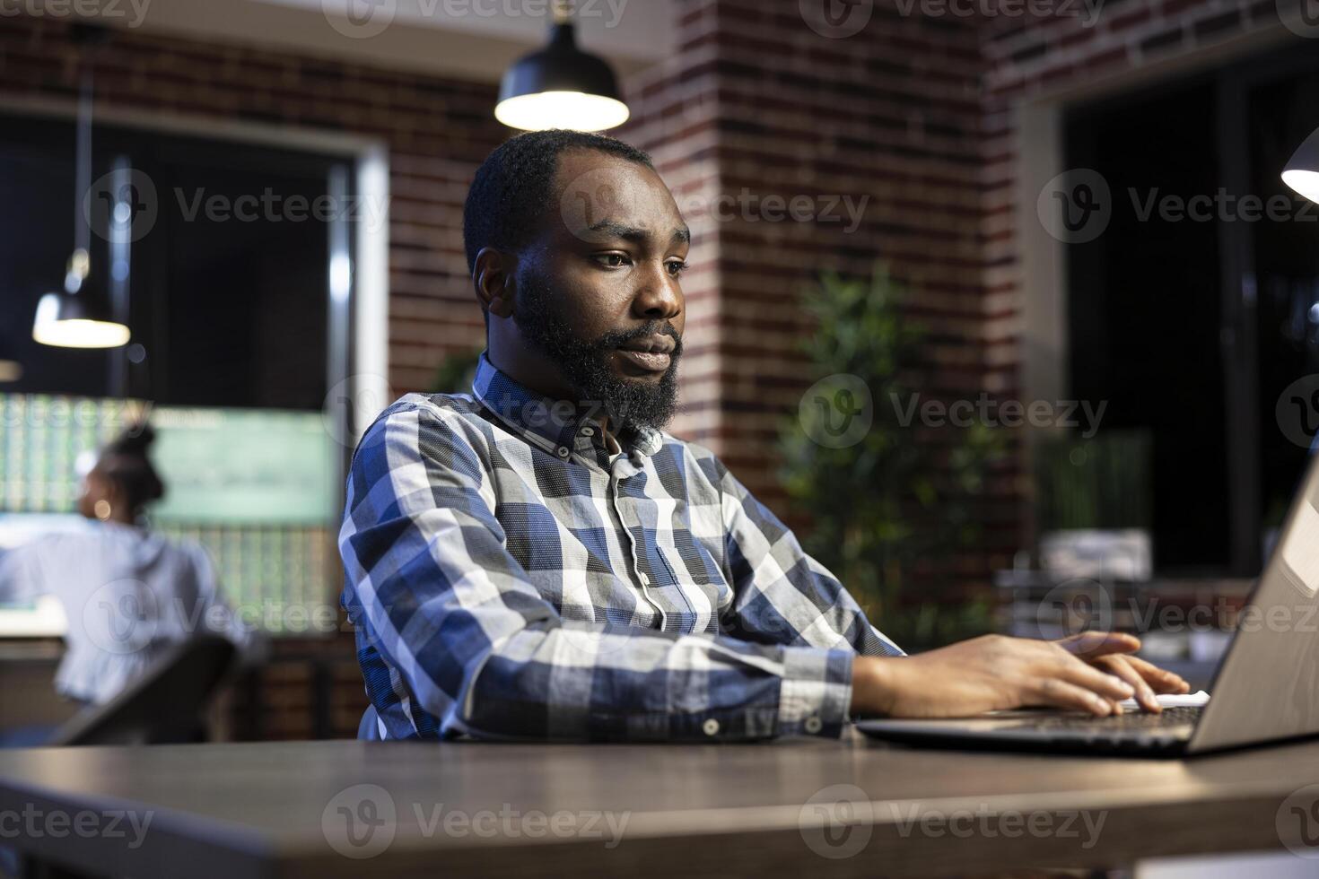 Male financial analyst works late at a brick wall office desk, reviewing documents and stock data on his laptop. The night setting creates a focused workspace for analyzing financial trends. photo