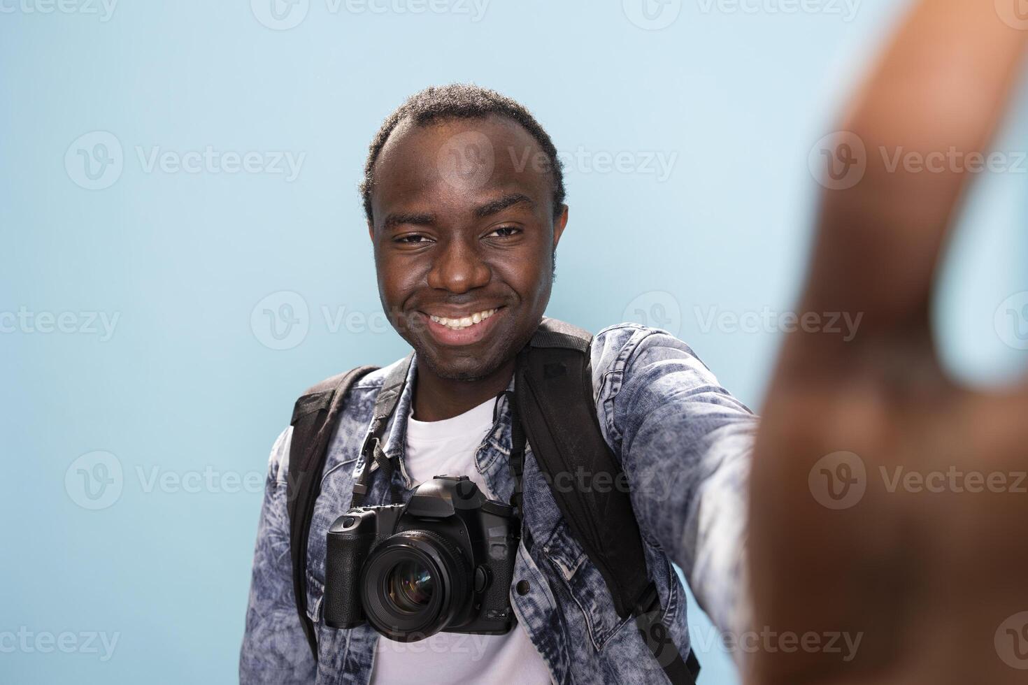 Smiling photographer recording content for vlogging channel in studio. POV of african american man with DSLR camera around his neck, cheerfully taking a selfie against isolated background. photo