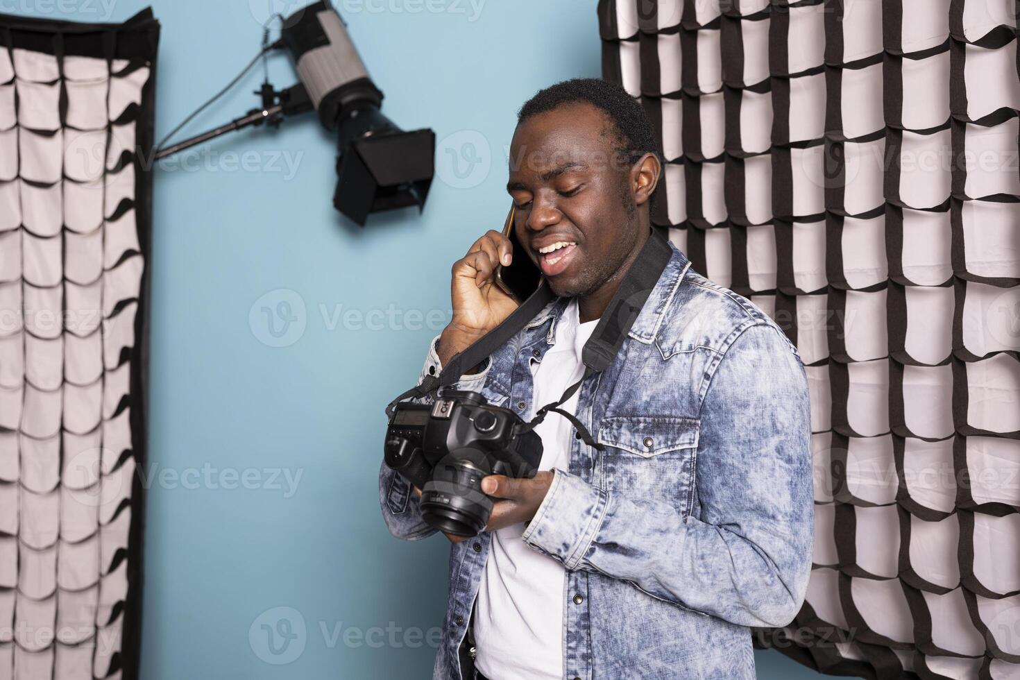 Black photographer on phone call, holding his DSLR and standing near his studio equipment. African American man with mobile device to his ear, wearing a camera around his neck and reviewing pictures. photo