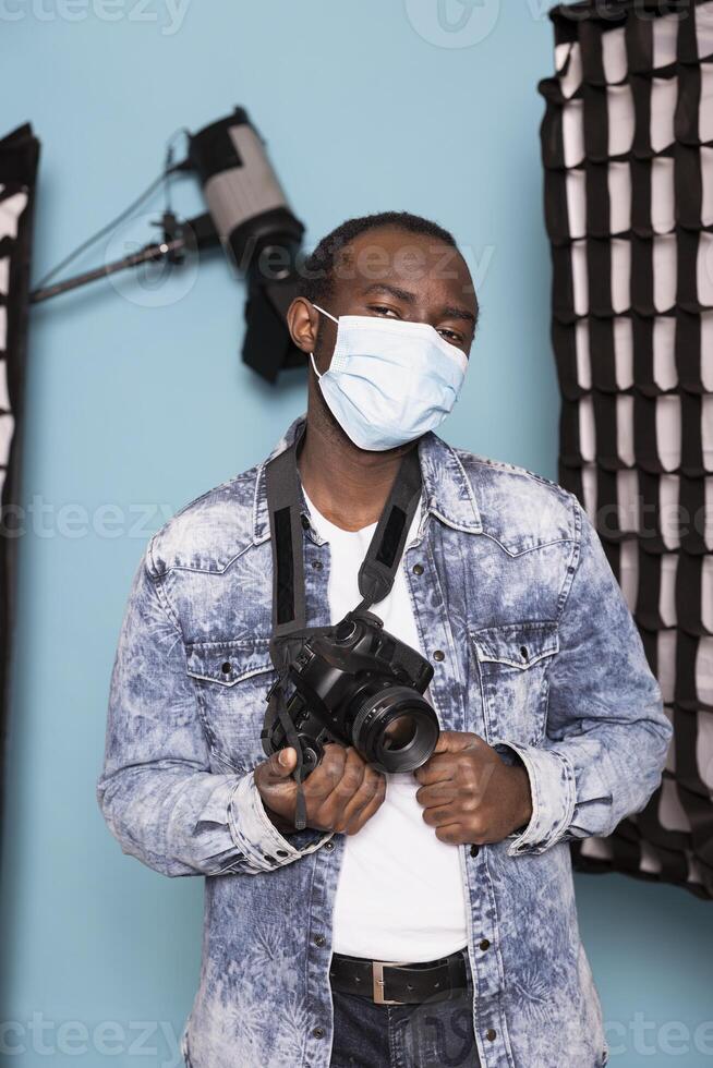 Portrait of a black cameraman protecting himself with a face mask while standing in blue background studio. African american male photographer with his nose and mouth covered, holding his dslr camera. photo