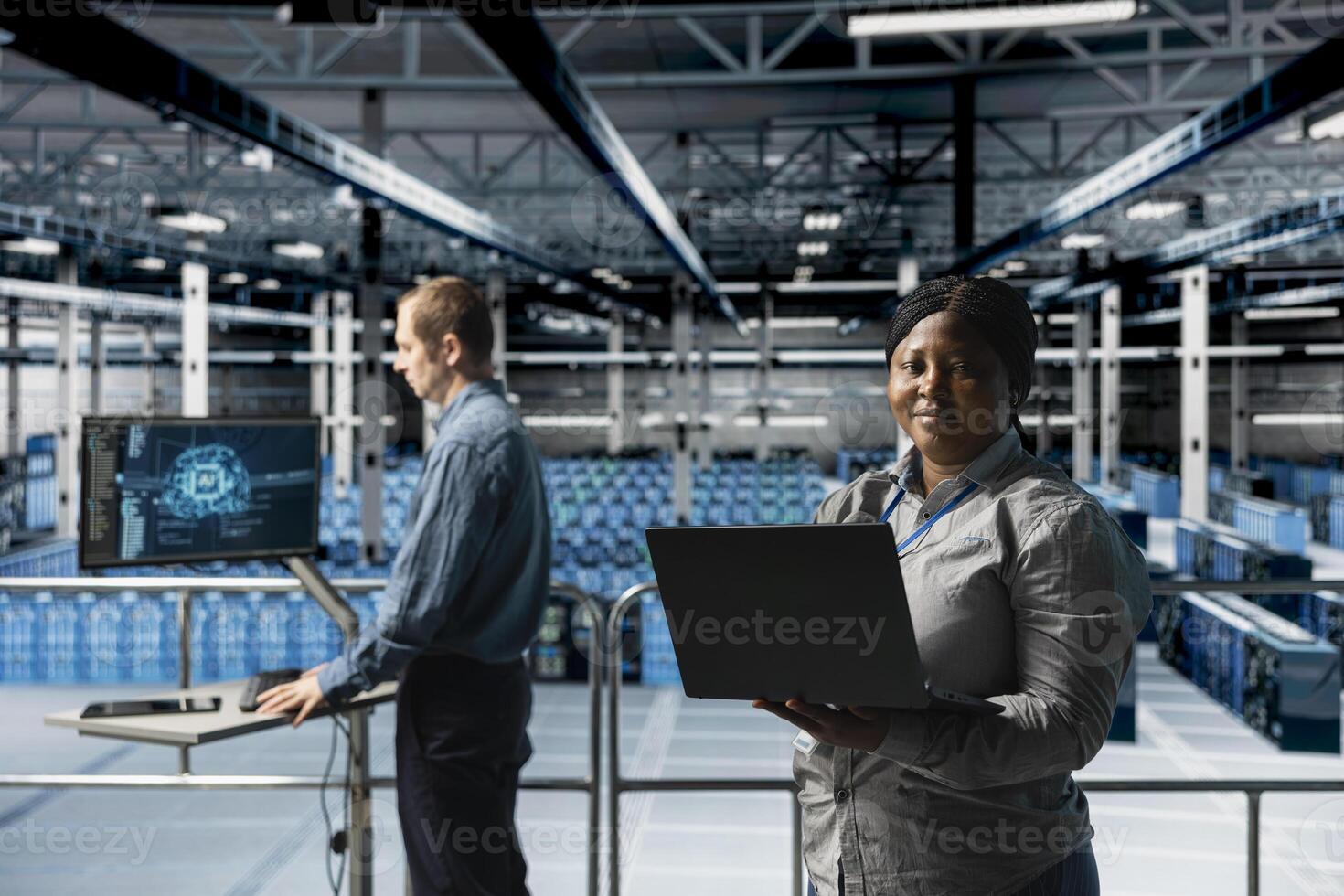 Portrait of smiling female engineer in server farm next to colleague doing infrastructure AI automatization. Happy african american woman in data center doing maintenance using artificial intelligence photo