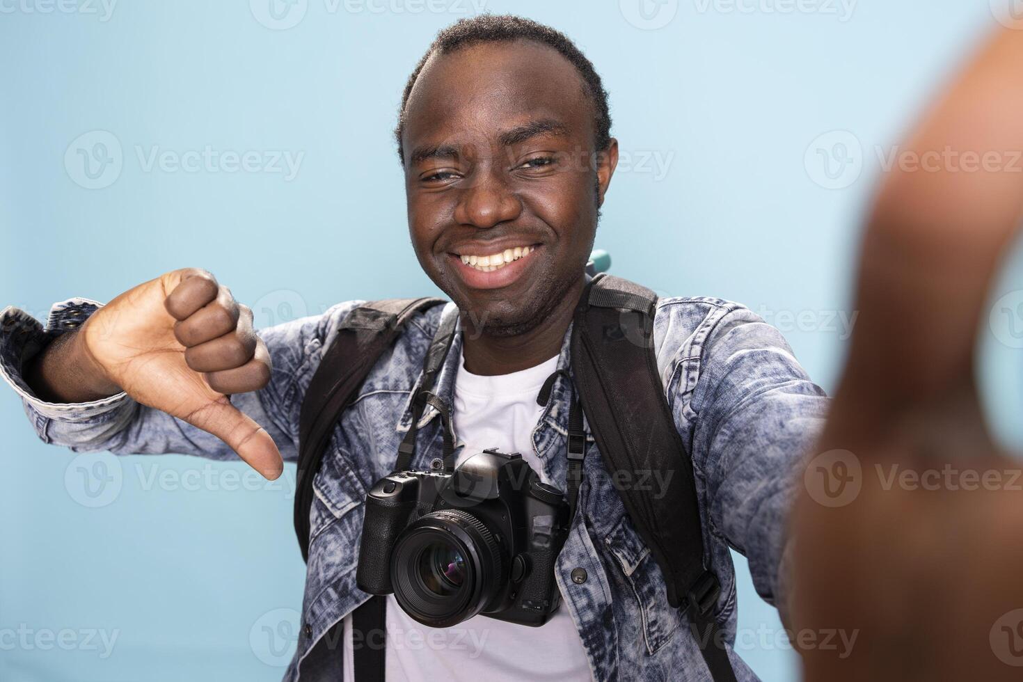 POV of male photographer standing and playfully giving a thumbs down against isolated background. African american vlogger with dslr camera and backpack recording himself as he gestures disagreement. photo