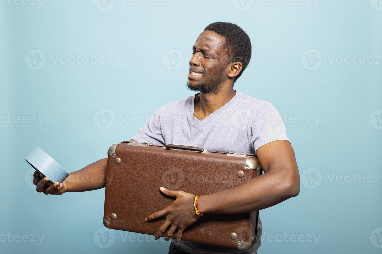 Male tourist looking at the clock, confused about the time as he carries his brown suitcase. Young man with worried expression, grasping both his luggage and alarm clock, afraid of missing his flight. photo