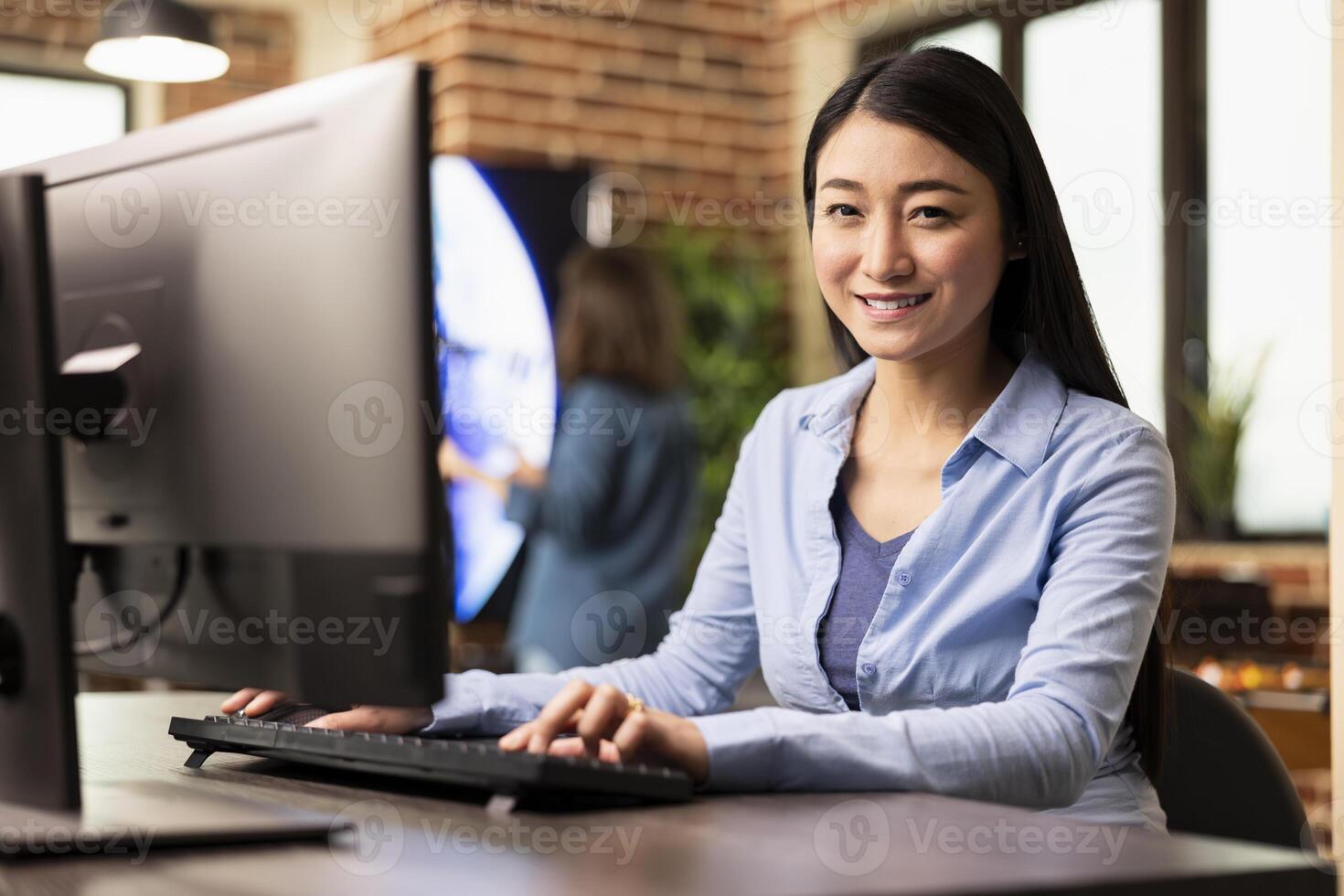 Young female employee sitting at desk in modern office, happily working on computer and looking at camera. Portrait of asian businesswoman typing on keyboard, engaged in work at creative workspace. photo