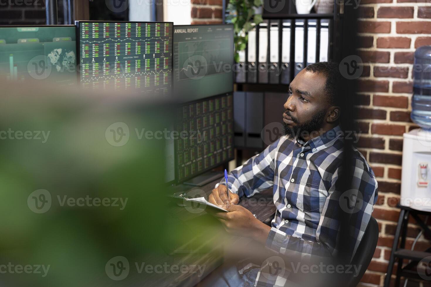 Male forex trader seated at desk, looking at screens displaying live trading session and taking notes in brick wall office. Financial expert with pen and clipboard, reviewing stock growth statistics. photo