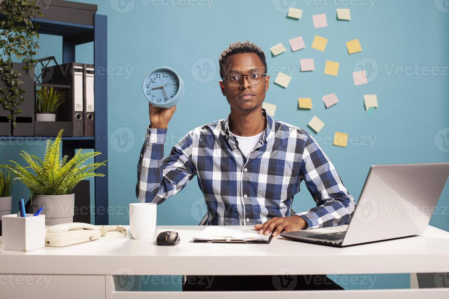 Portrait of a young African American freelancer sitting at office desk with a clipboard and showing an analogue clock. Black male business analyst managing paperwork and presenting the time. photo