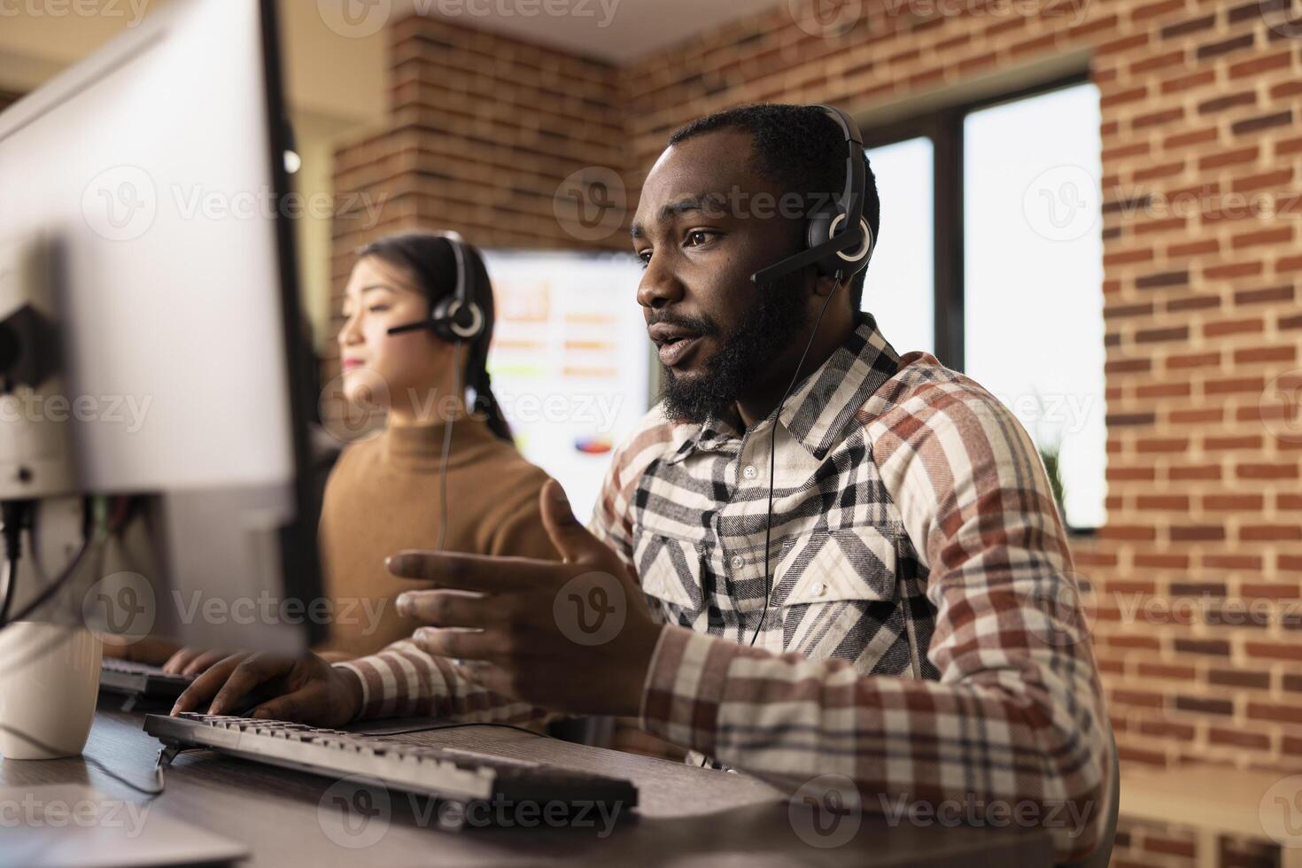 African American man wearing wired headphones, providing customer support while reviewing information on computer screen. Black male employee speaking into headset microphone, assisting online client. photo