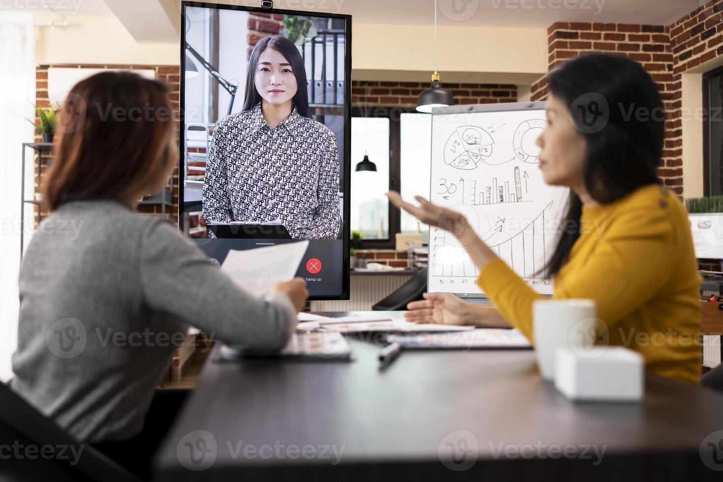 Company advisors seated at desk with documents, shares business updates with a remote client via call on led screen. Asian women discussing project analysis during virtual meeting with manager. photo