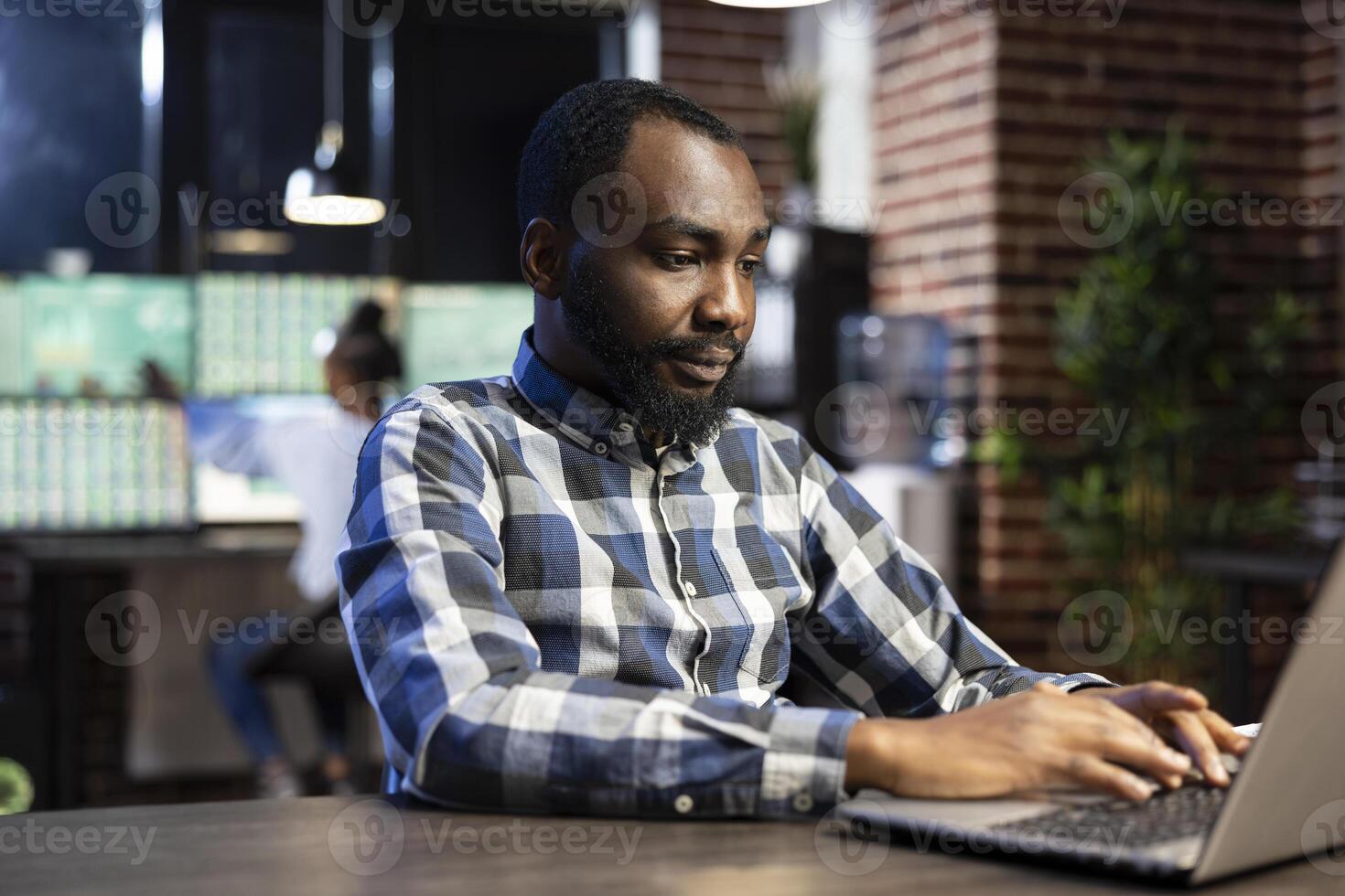 Black male manager seated at desk, using a laptop and reviewing financial online documents. The office features a brick wall and a calm night shift ambiance, enhancing the professional workspace. photo