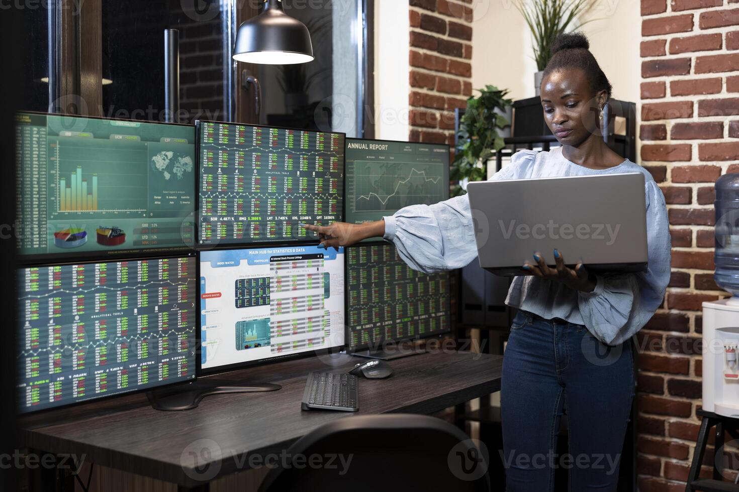 African American stockbroker standing by desk, comparing forex market data on laptop and several monitors in professional nighttime trading environment. Black woman woman working in tech savvy office. photo