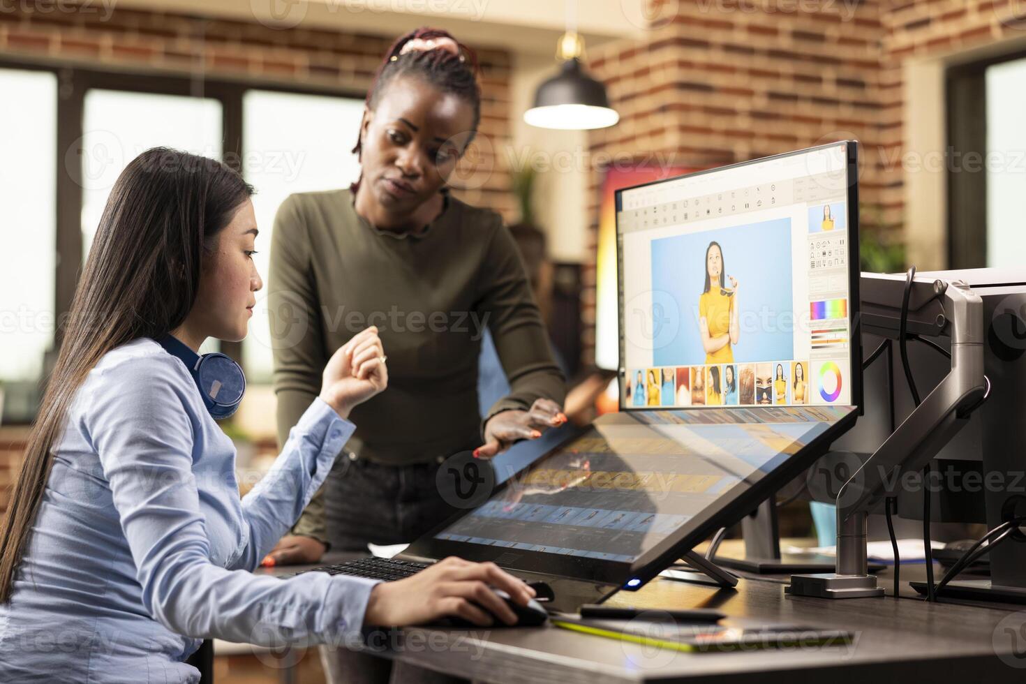 Multiethnic designers collaborate on photo retouching in studio, refining images for a project. In creative workspace, diverse women carefully select and color grade images for a commercial campaign.