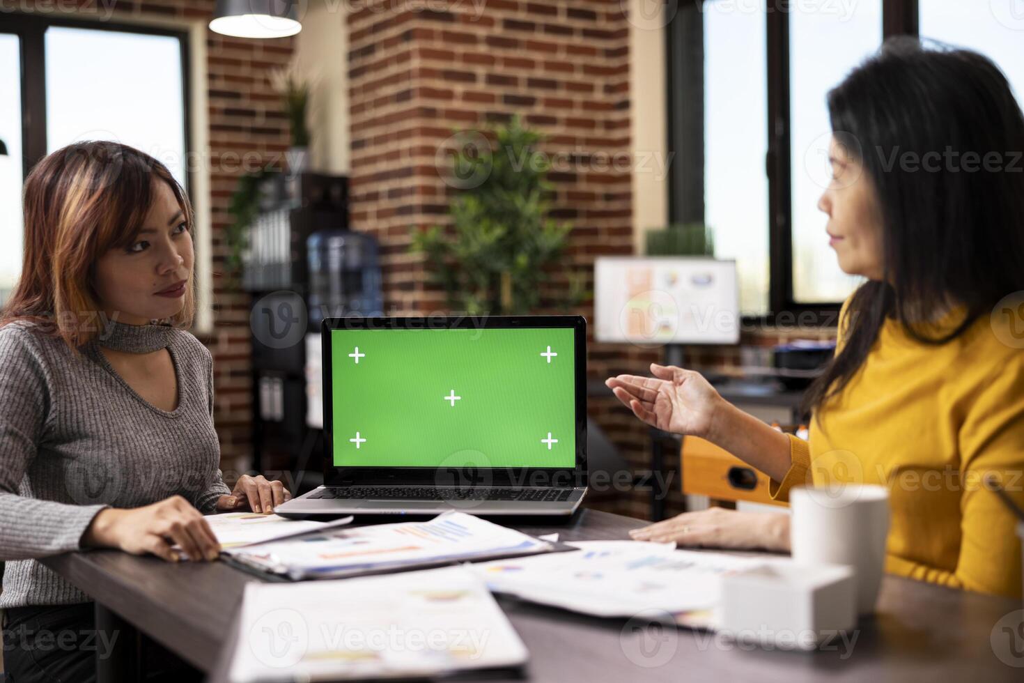 Digital device displaying a blank chromakey template sits on the table alongside scattered paperwork, with two women having a business discussion. Strategy meeting between coworkers. photo