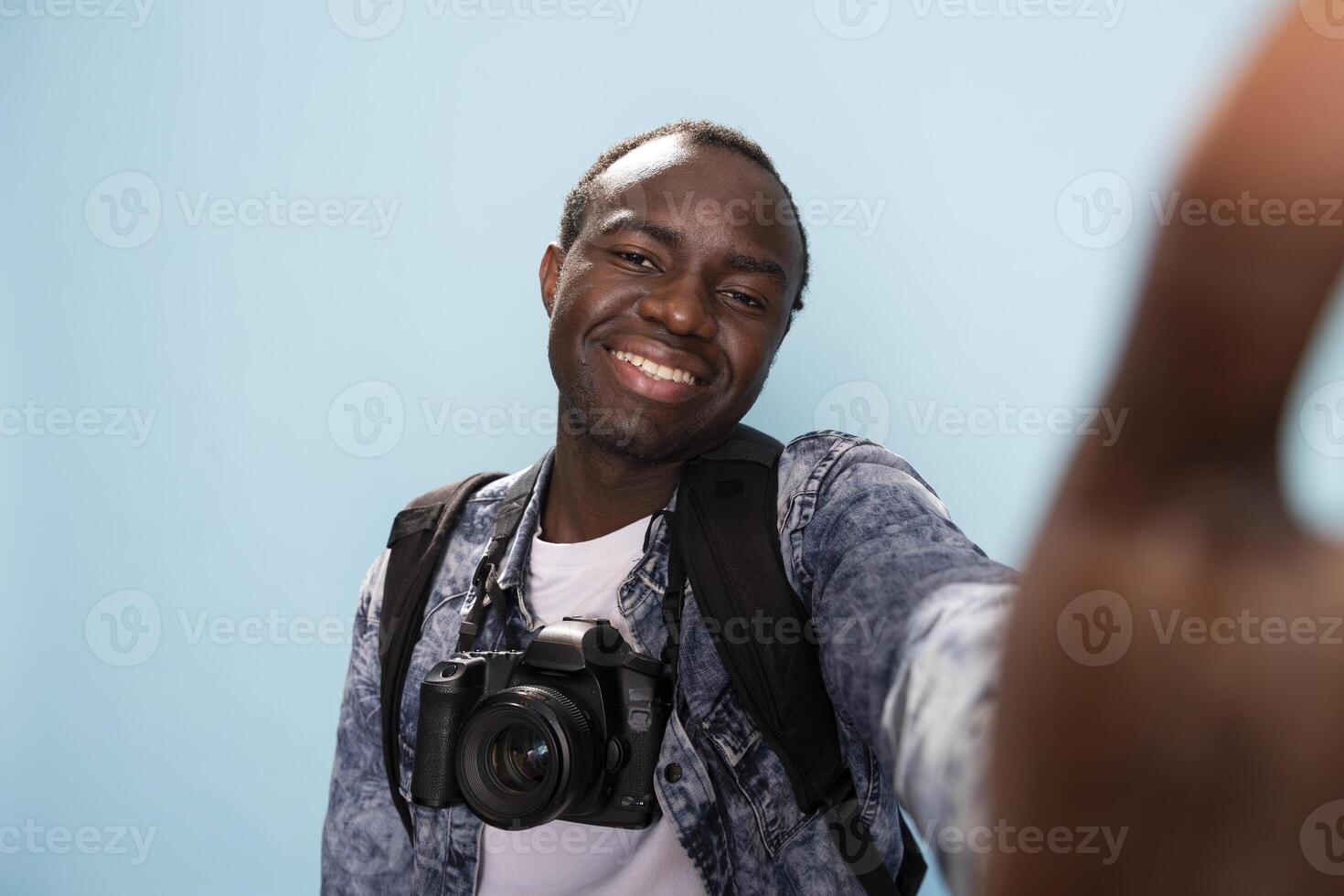 Male photographer using a camera to record himself in a blue background studio. POV of African American man with a DSLR around his neck, smiling towards the digital device in his hand. photo