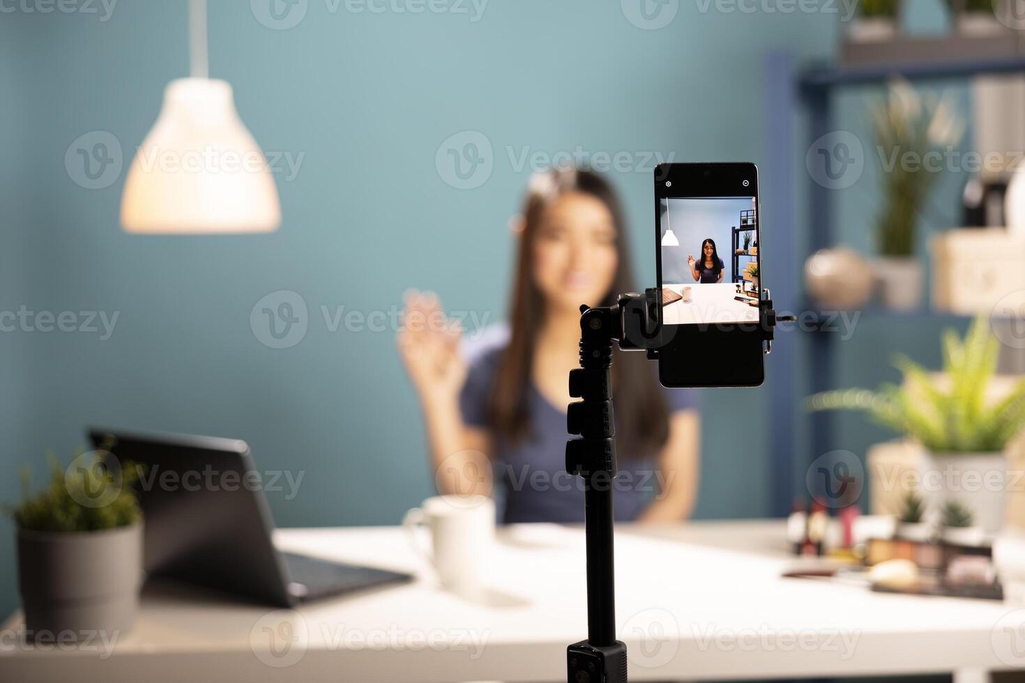 Closeup of a mobile device focused on a beauty influencer in the screen display. Asian woman waving at phone camera, ready to demonstrate makeup techniques to her vlogging channel. photo