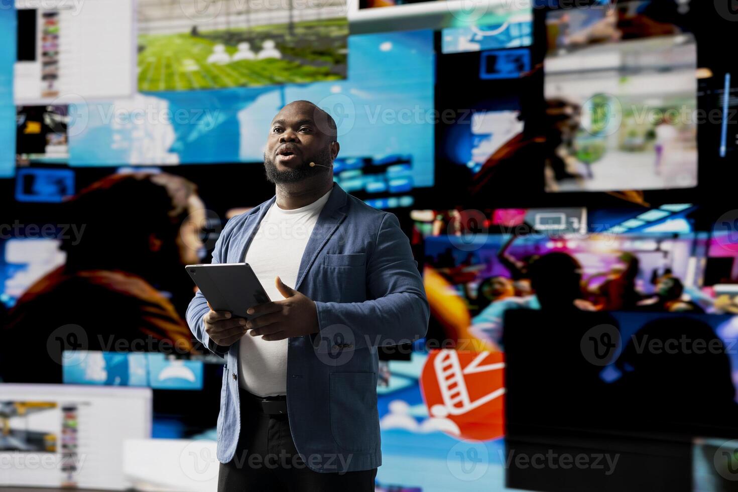Black presenter hosting a conference on stage surrounded by media visuals, symbolizing a global convention with live audience to discuss about online trends and mobile apps. Public event. photo