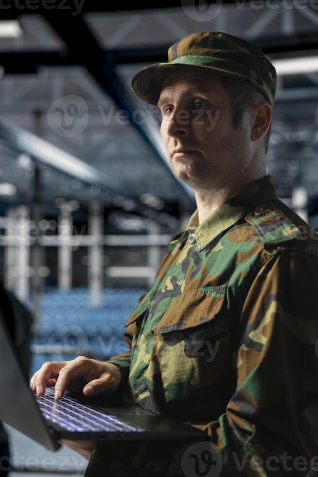 Military officer managing logistics operations using laptop in server room command center. Soldier wearing uniform running diagnostics on infrastructure in army data center photo