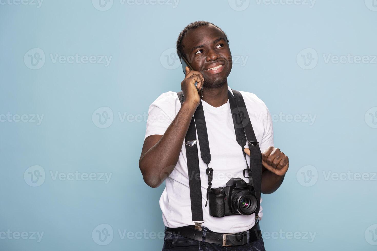 Black man in casual clothing with suspenders, holding his phone to his ear and wearing a camera around his neck. He stands in a casual pose, blending creativity with a tech savvy lifestyle. photo