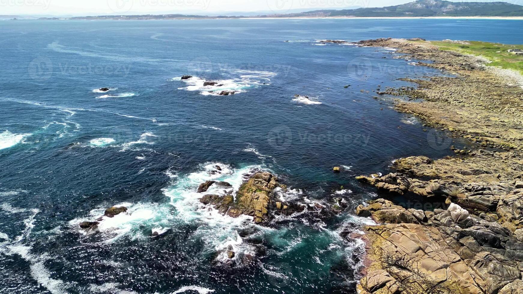An aerial view shows a rocky coastline with waves crashing beneath blue skies and pristine clear waters photo