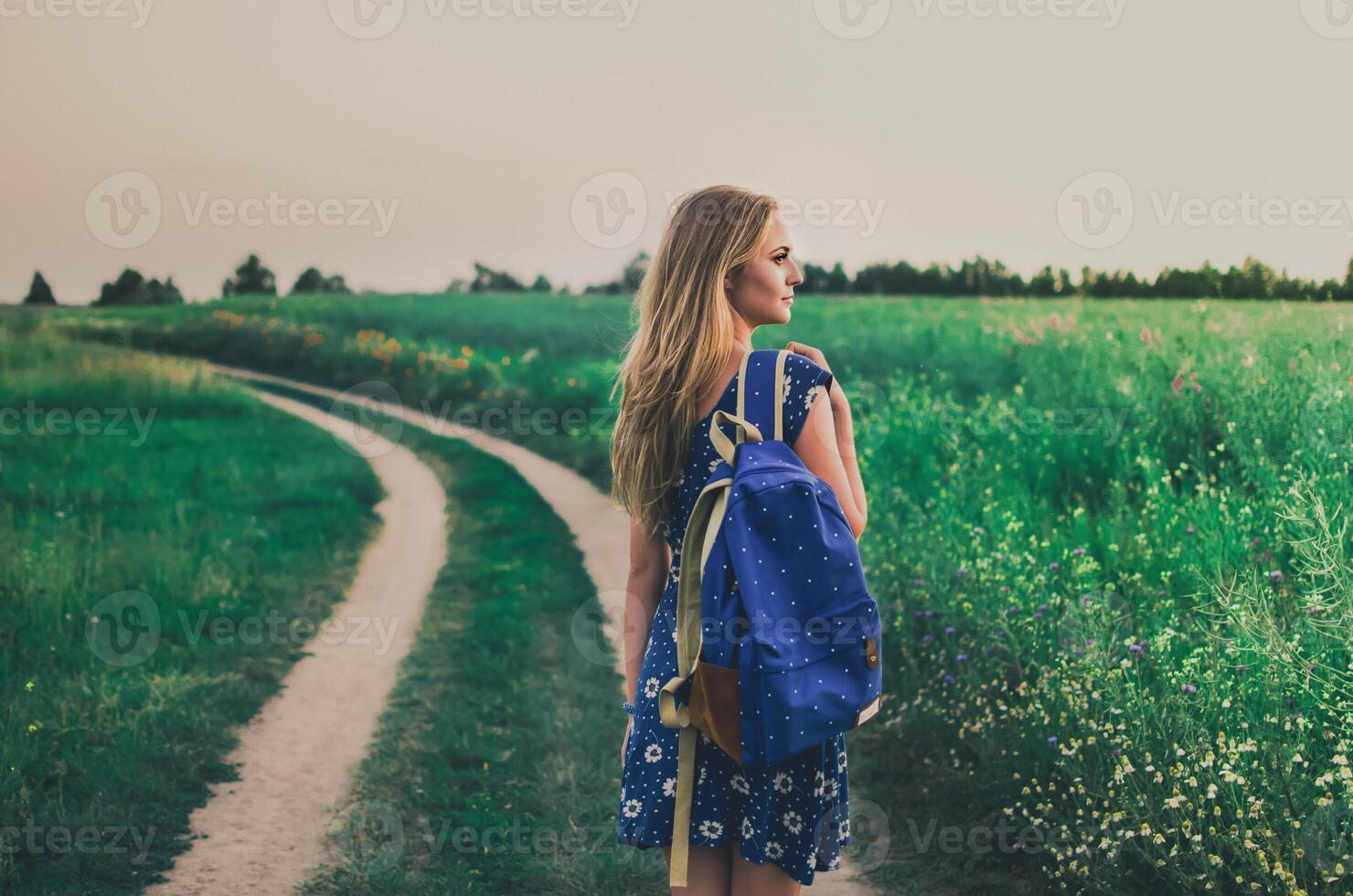 woman stands in a dress and a backpack in middle of a green field On a walking path photo