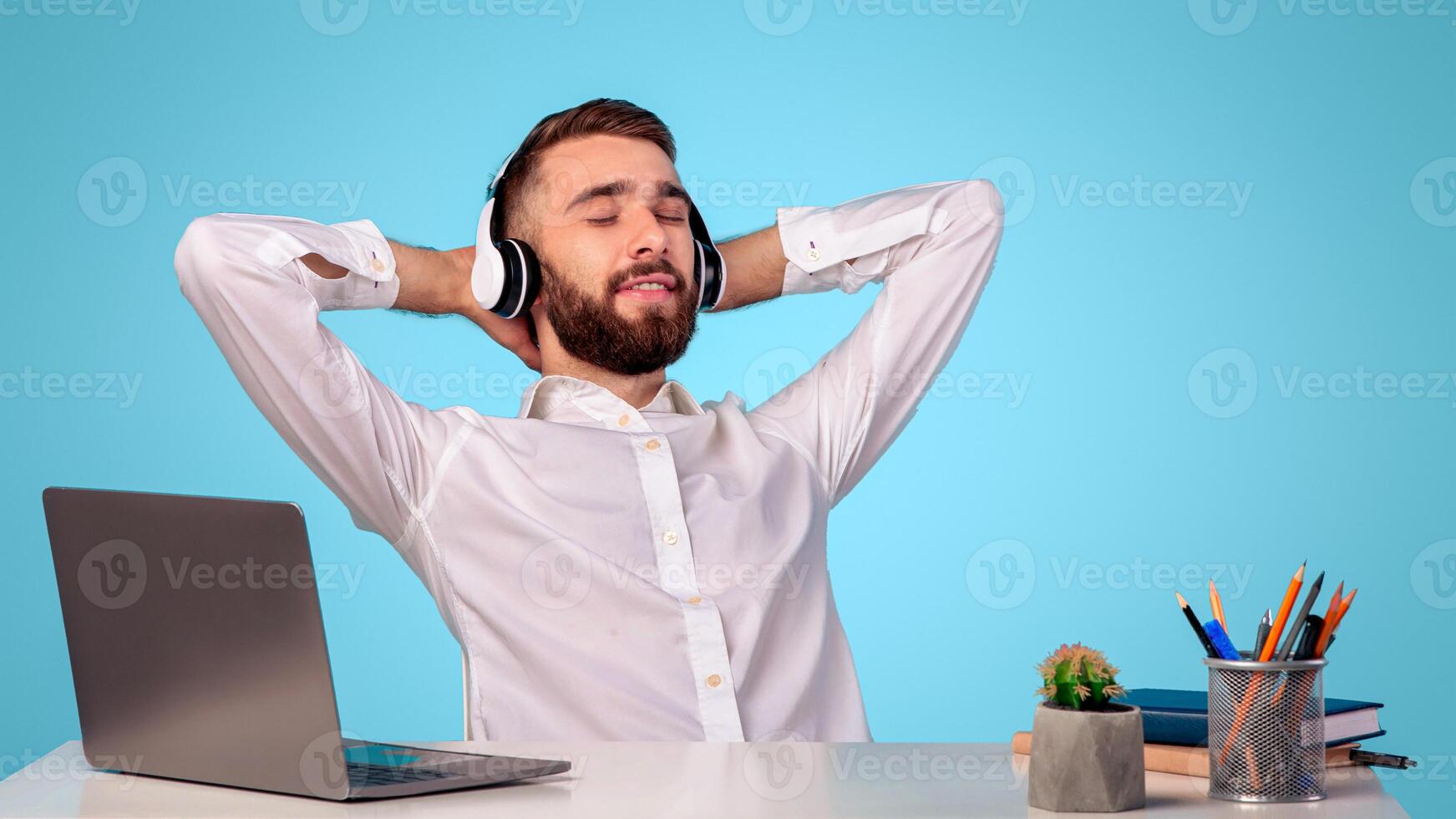 Taking break from work. Smiling Caucasian man listening to music in headphones at his desk on blue studio background. Relaxed young guy sitting at table with laptop and enjoying great song photo