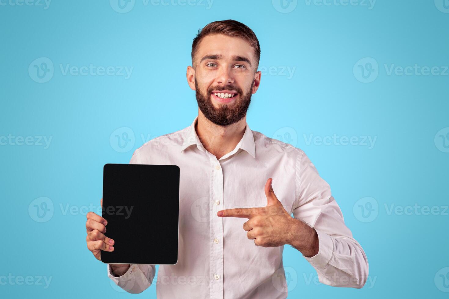 Young businessman pointing at tablet with empty screen on blue studio background, mockup for application or website design. Cheerful millennial guy showing touch pad with space for your ad photo