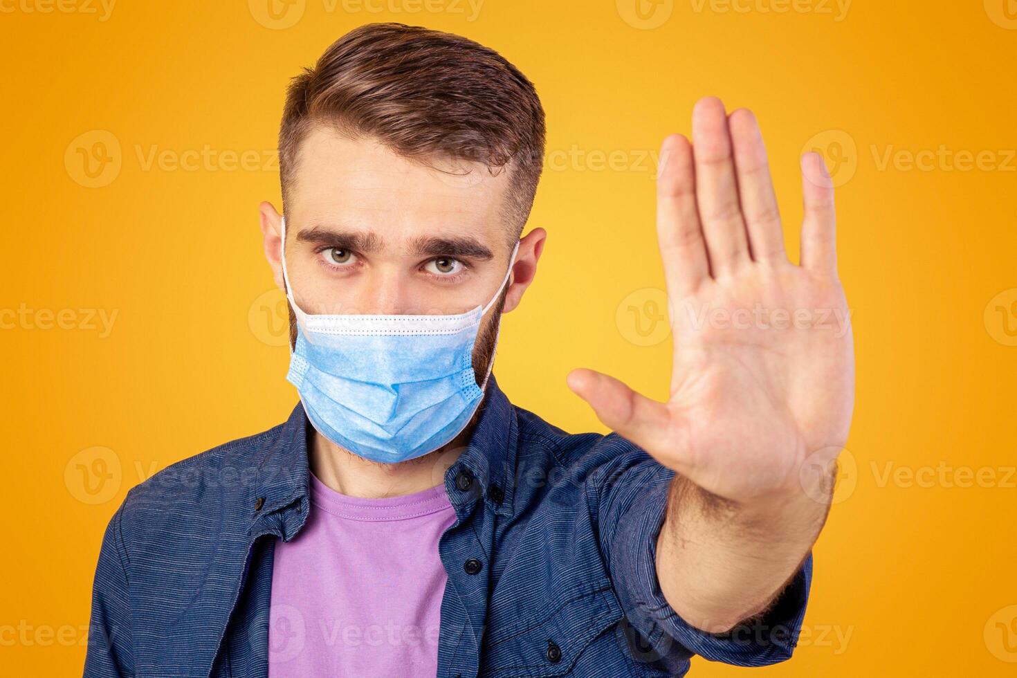 No to coronavirus. Young Caucasian man in medical mask making STOP gesture on orange studio background. Millennial guy wearing protection, expressing negative attitude to worldwide epidemic photo