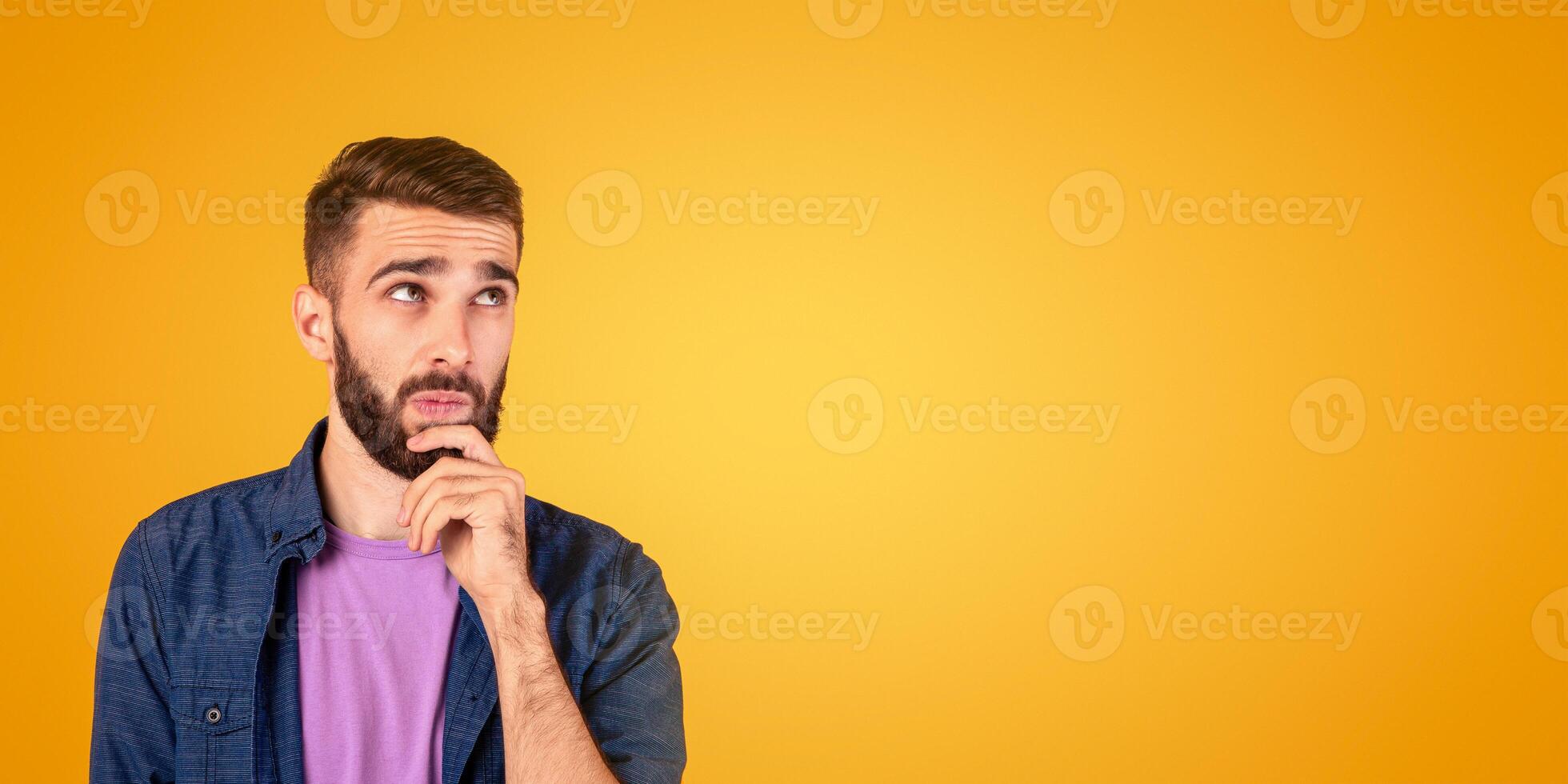 Portrait of pensive young bearded man touching his chin, deep in thought over orange studio background. Doubtful millennial guy thinking and making decision, dreaming about something photo