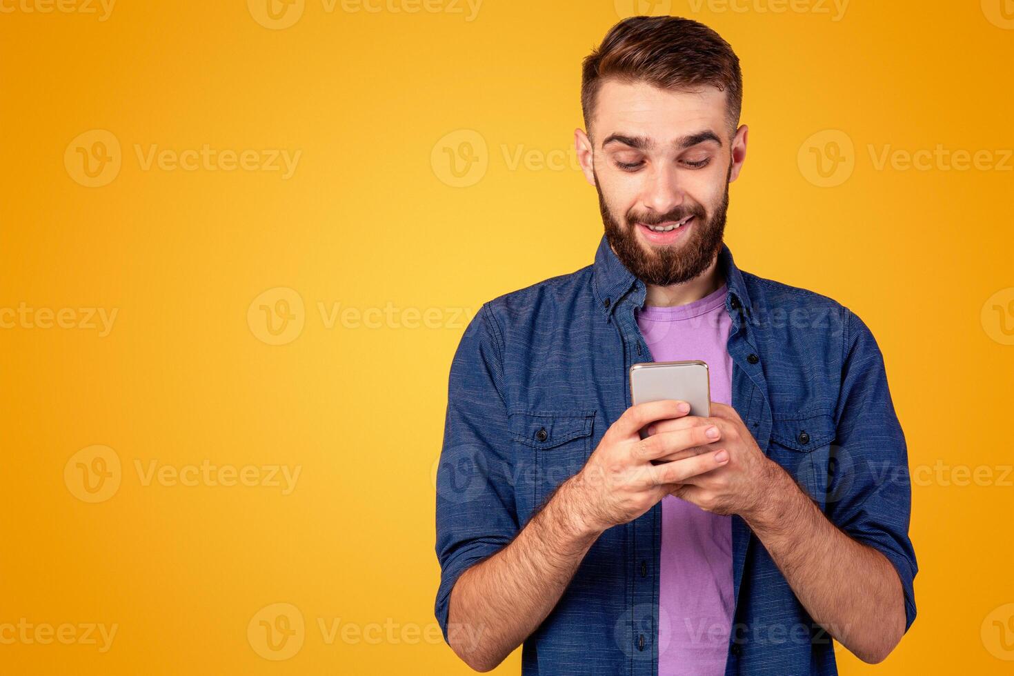 Handsome bearded guy with smartphone browsing internet, communicating online or shopping over orange background. Young man working or studying remotely using mobile device photo