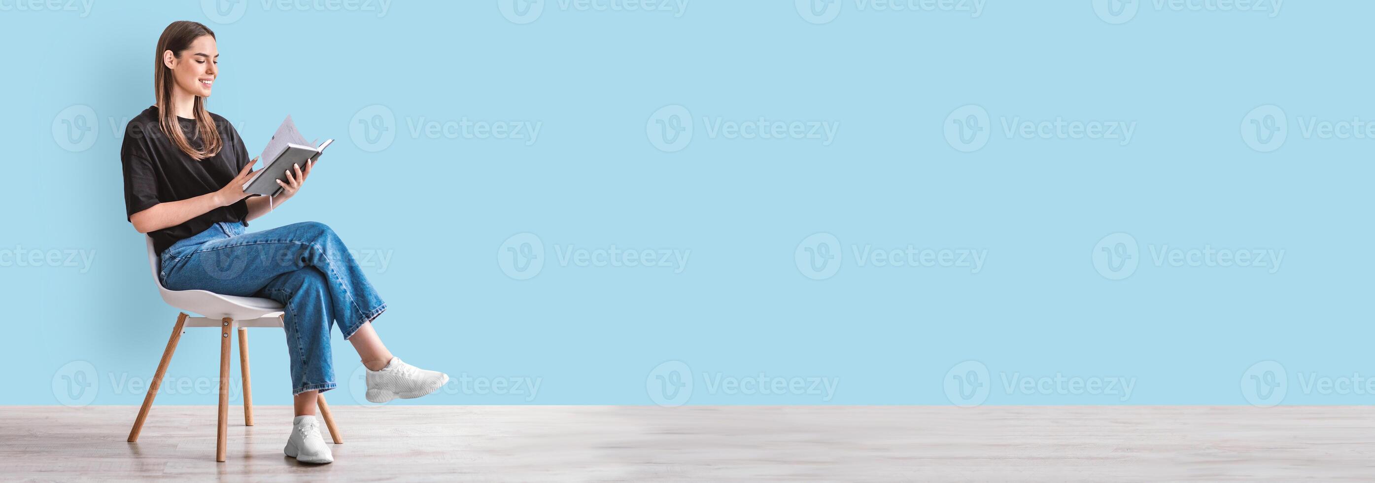 Hobbies and activities. Happy young caucasian woman reading book, sitting on chair, blue studio wall, panorama with copy space. Millennial lady enjoying interesting story, studying for exam, mockup photo