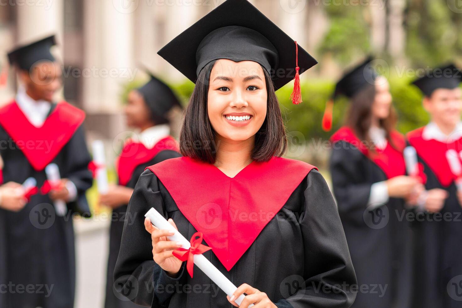 Happy young asian woman student cheerfully posing while graduation ceremony, wearing graduation robe and hat, holding diploma and smiling at camera, international group of students chatting on background photo