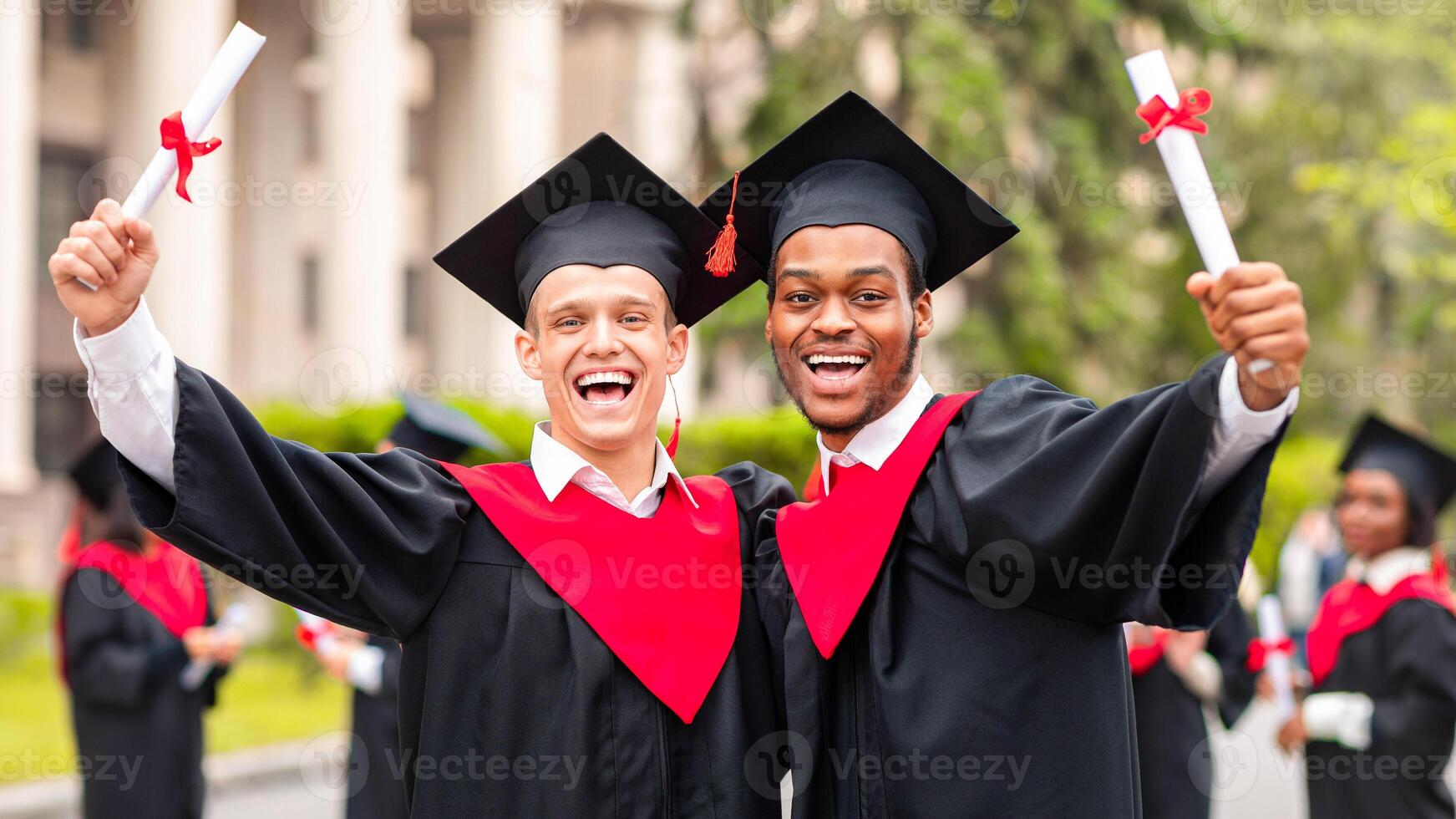 Happy guys students posing together next to university, wearing graduation costumes and showing diplomas. Cheerful multiracial group of male friends celebrating graduation, portrait photo