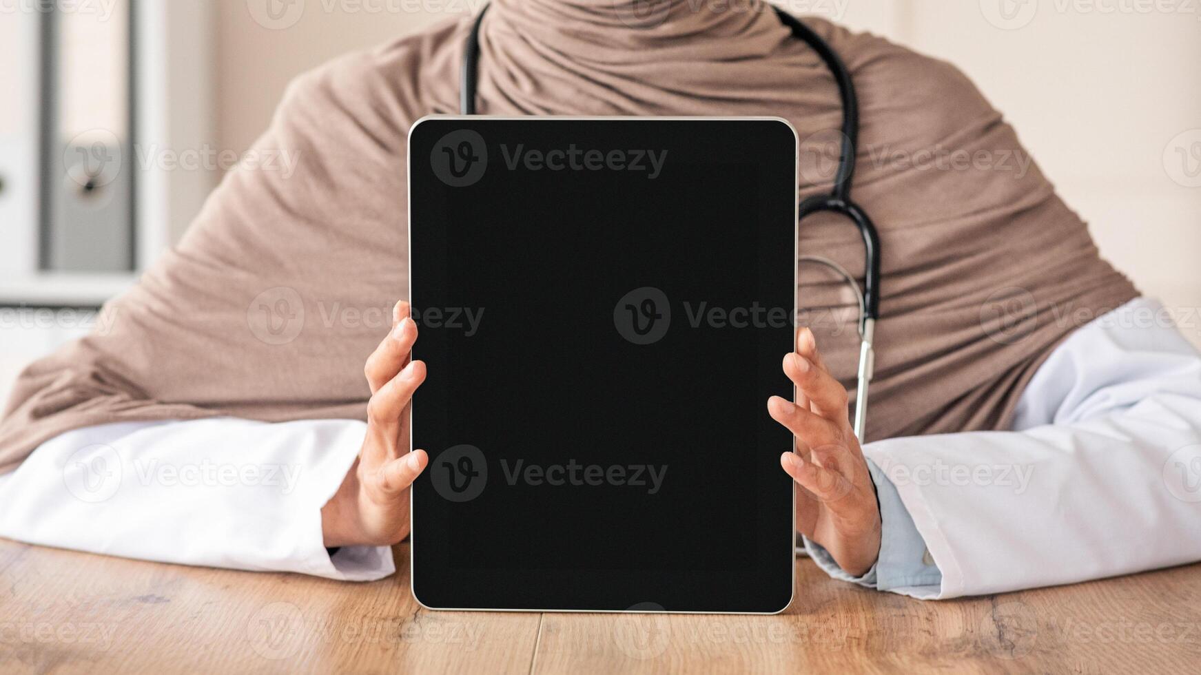 Cropped of female doctor muslim woman in hijab showing digital tablet with blank screen, mockup, copy space. Unrecognizable doctor using modern technologies in her job, working and training online photo