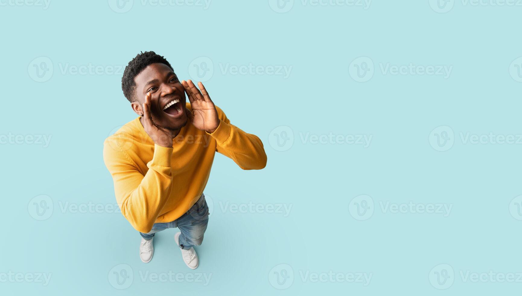 Cheerful african american guy posing over blue studio background, holding hands to mouth and saying something out loud, panorama with copy space. Handsome young black man making announcement, top view photo
