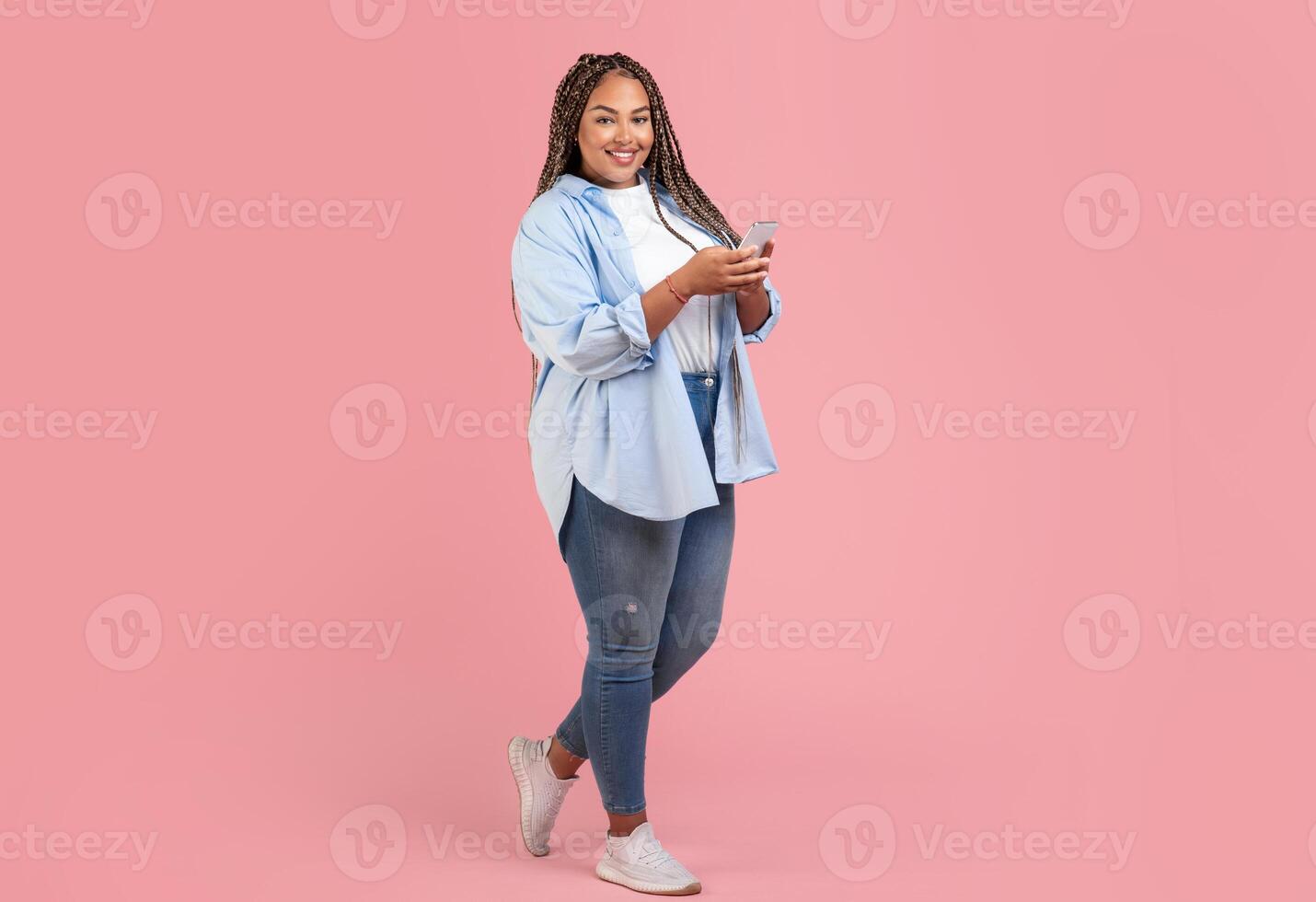 Black Plus Size Woman Using Smartphone Standing On Pink Studio Background, Smiling To Camera. Cheerful Female Using New Mobile Application And Texting. Technology And Gadgets. Full Length photo