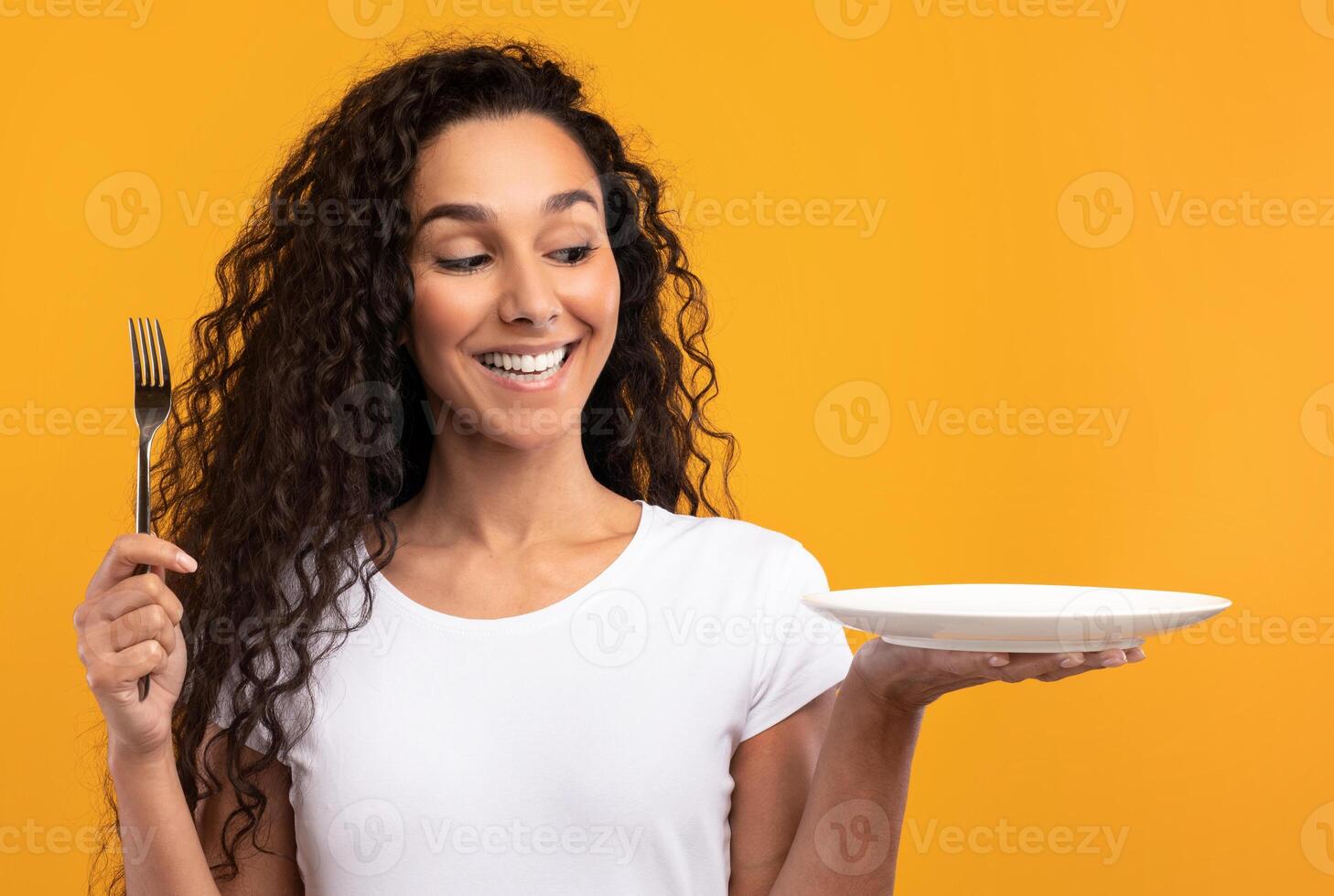 Portrait of excited hungry young lady holding and showing fork and empty plate in hand, funny young woman wants to eat posing isolated on yellow orange studio background, free copy space, mockup place photo