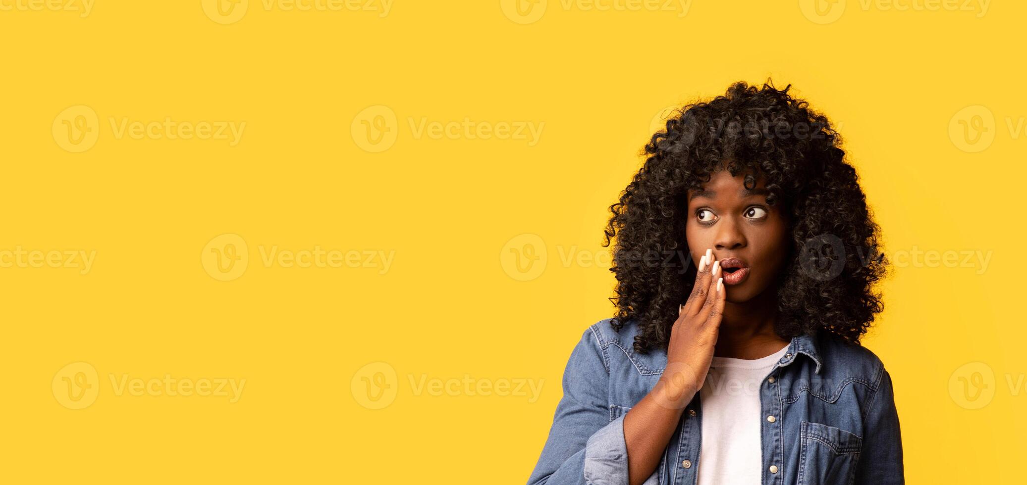 Amazed african american young woman looking at empty space for advertisement or announcement, touching her face. Pretty curly black lady in jeans shirt looking at copy space over yellow background photo