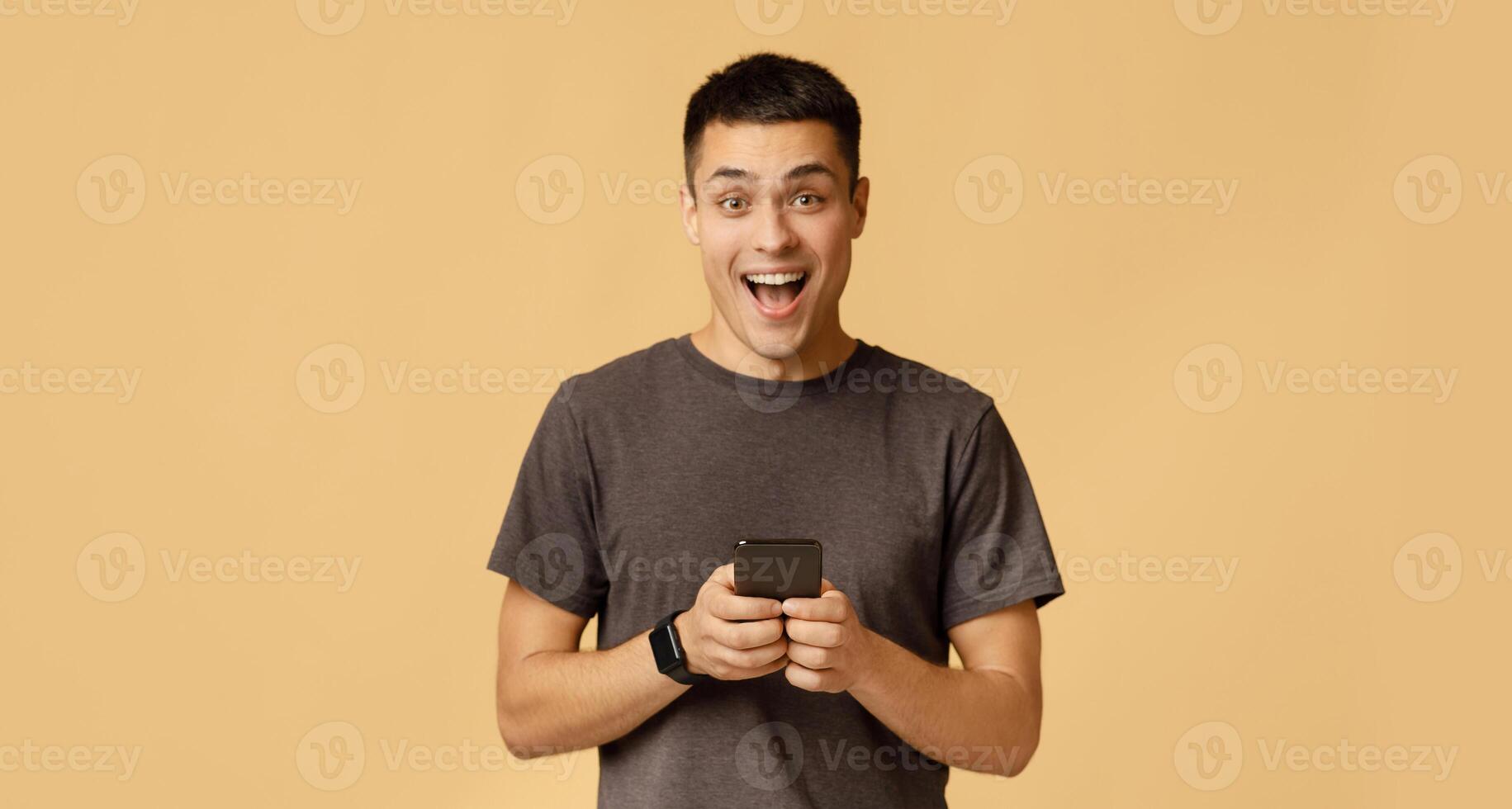 Emotions of success, victory, cashback and great message about win. Happy millennial guy student in brown t-shirt shouts with joy and holds smartphone, isolated on beige background, studio shot photo