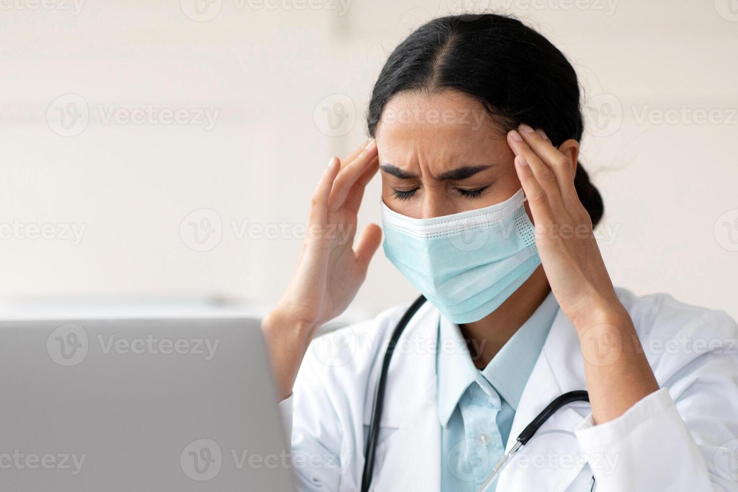 Female doctor young brunette woman in protective face mask having headache while working with laptop at clinic, sitting at workdesk with closed eyes, touching her temples, copy space photo