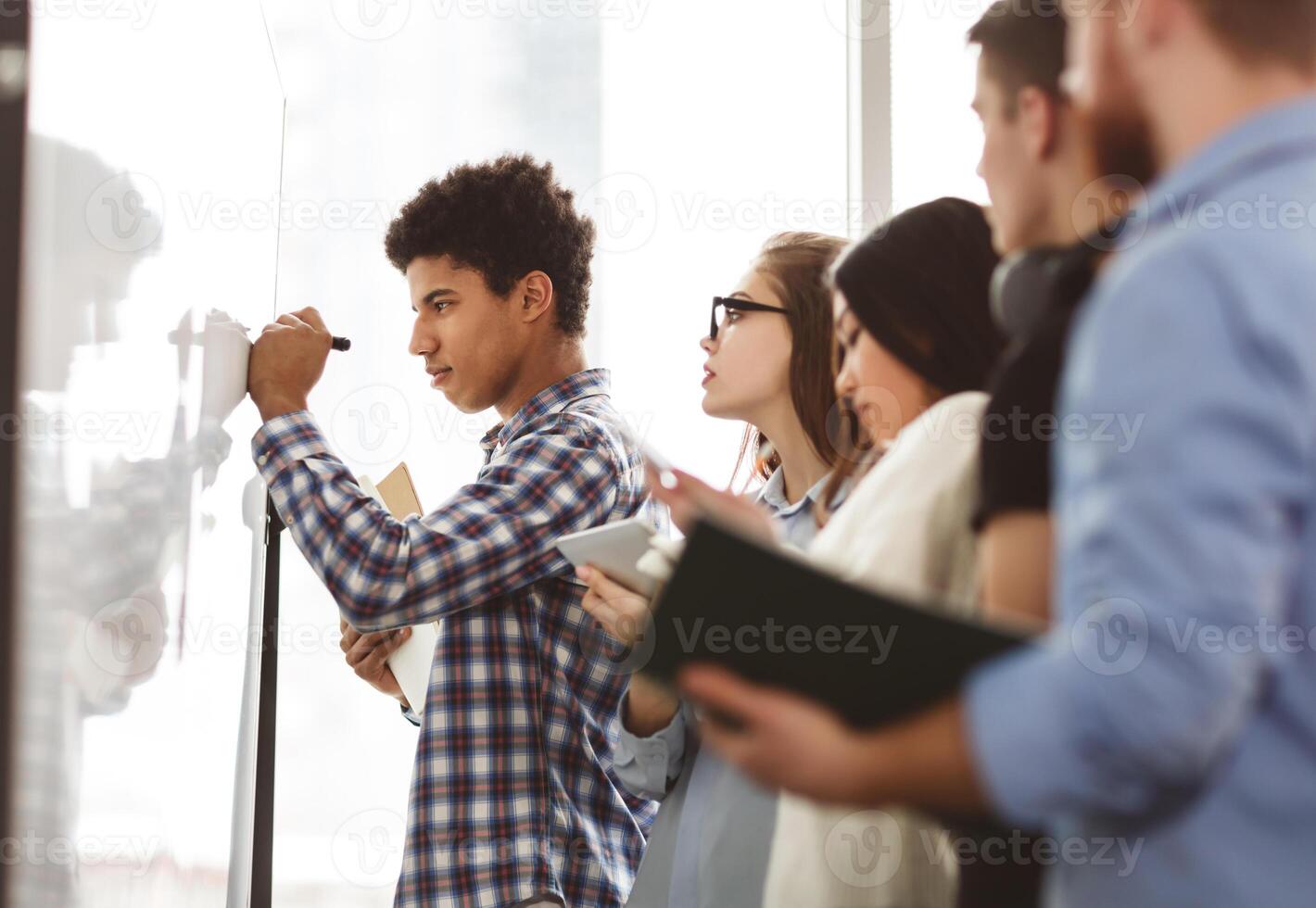 Afro guy writing algebra equations on whiteboard in classroom, friends checking answers photo