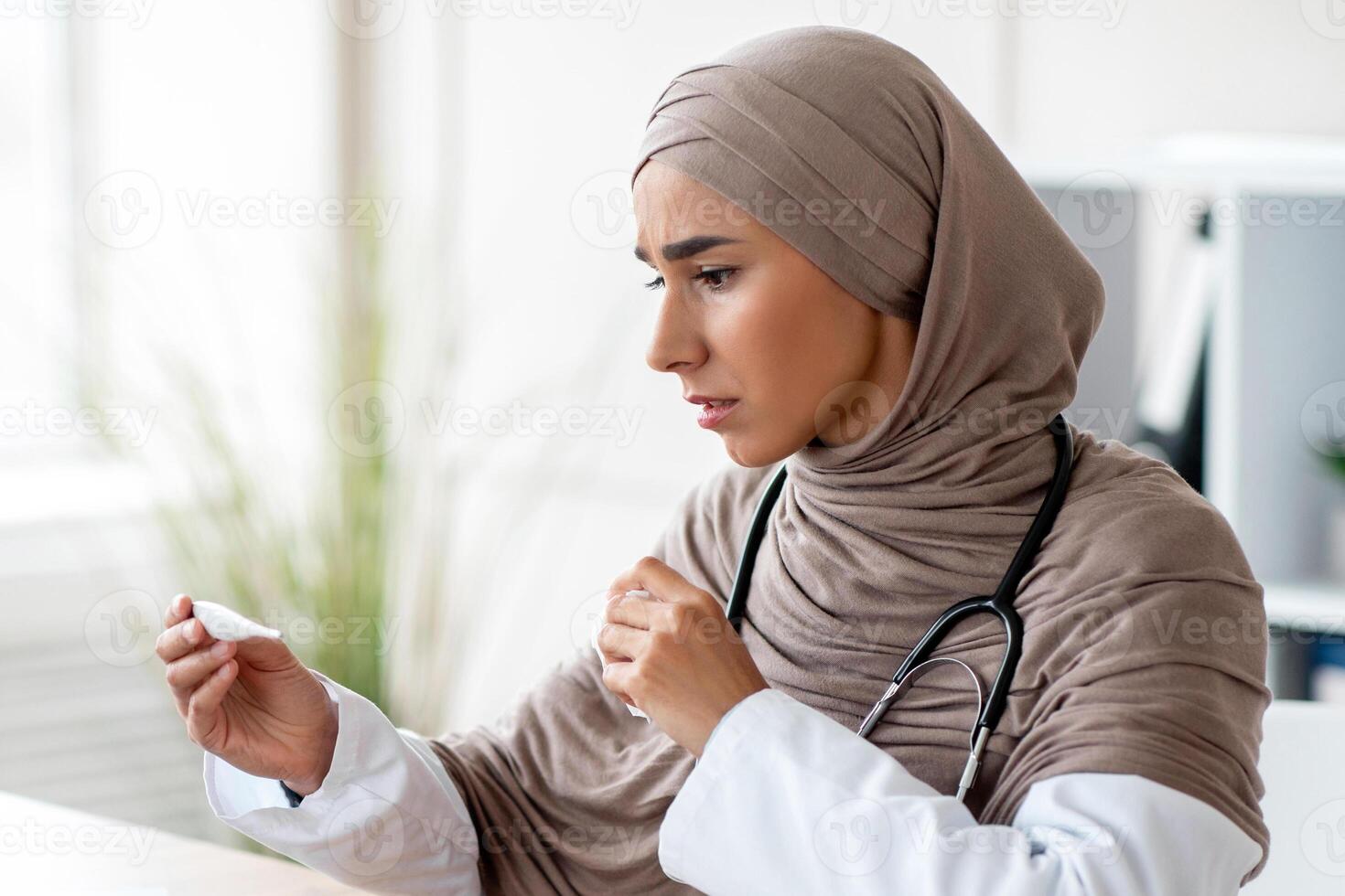 Sick young muslim woman in hijab doctor looking at thermometer, checking fever, sitting at workdesk with laptop in office at clinic, got sick during COVID-19 pandemic, copy space photo