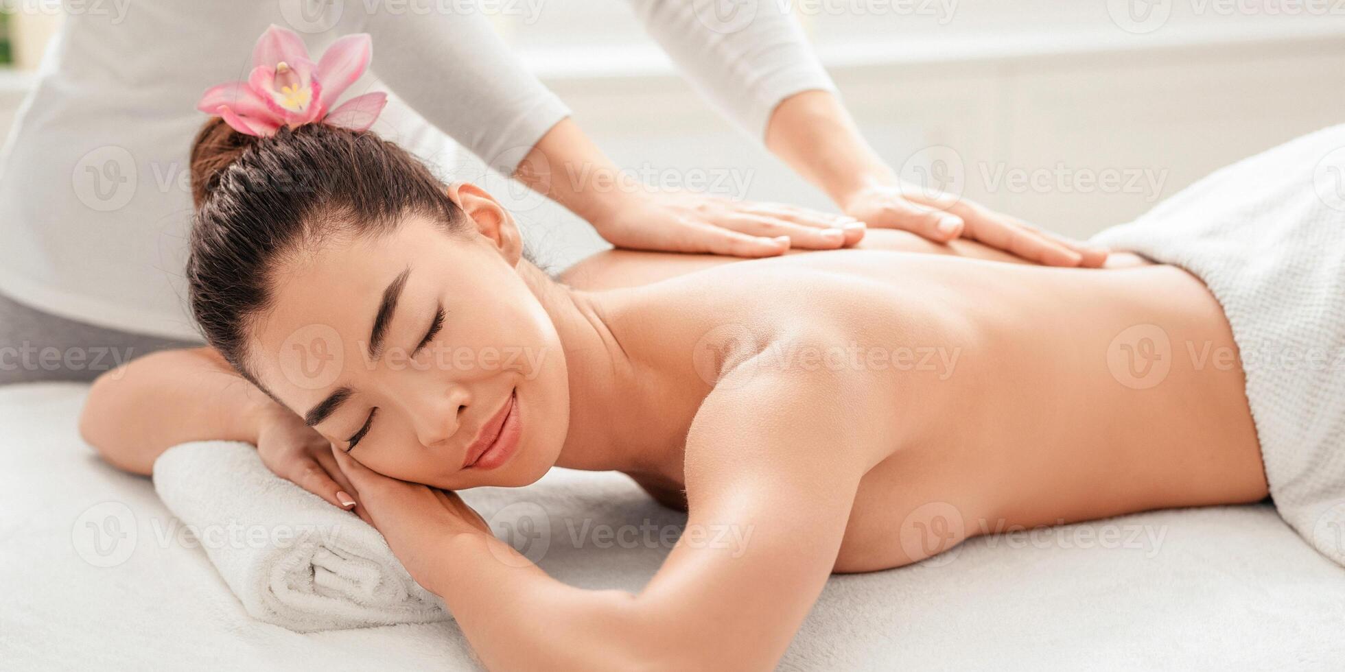 Enjoying massage. Beautiful asian lady relaxing during body treatment with professional masseur therapist in salon, lying on spa table with eyes closed, pampering herself with wellness procedures photo
