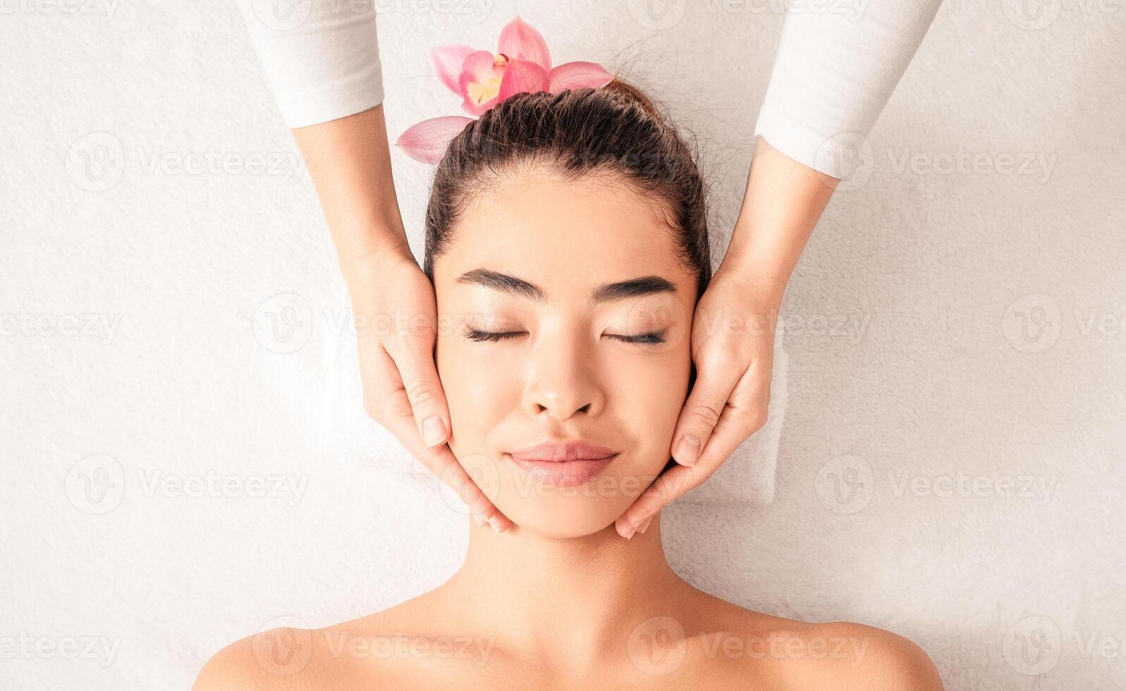 Young asian woman enjoying skin lifting face massage, getting professional beauty treatments at wellness center, attractive female relaxing on massage table with closed eyes, top view with copy space photo