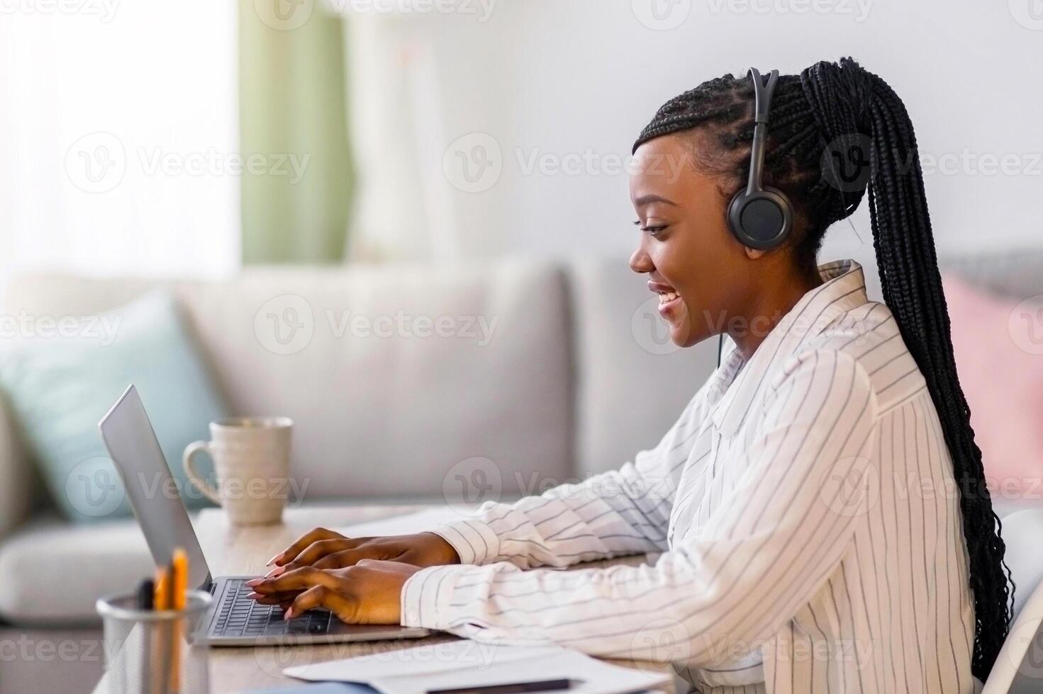 Positive young black woman with headphones sitting in front of laptop, typing on keyboard. female freelancer working from home during COVID-19 pandemic, copy space, side view photo