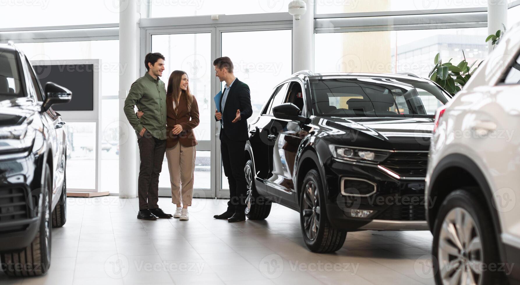 Car purchase or rental. Positive young spouses speaking to salesman about buying new auto at dealership, panorama. Successful millennial manager showing clients choice of vehicles at salon photo