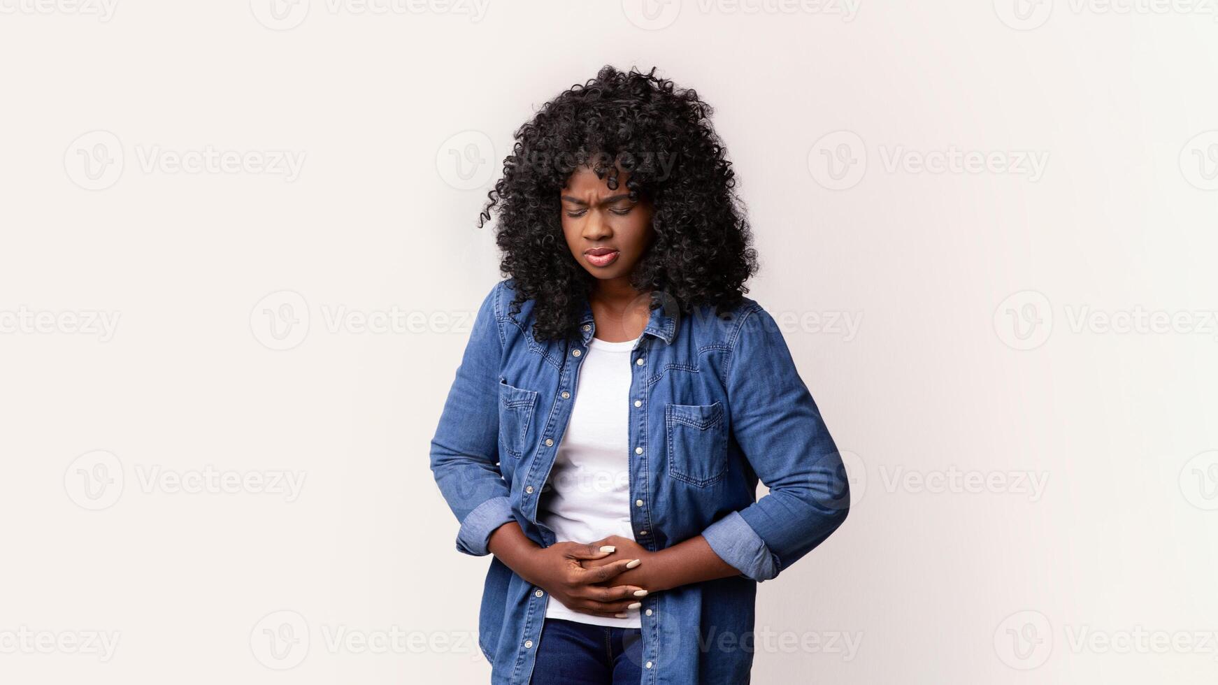 Sad black lady touching her belly on grey studio background, looking at copy space, panorama. Upset african american young woman suffering from abdominal pain, having menstruation cramps photo
