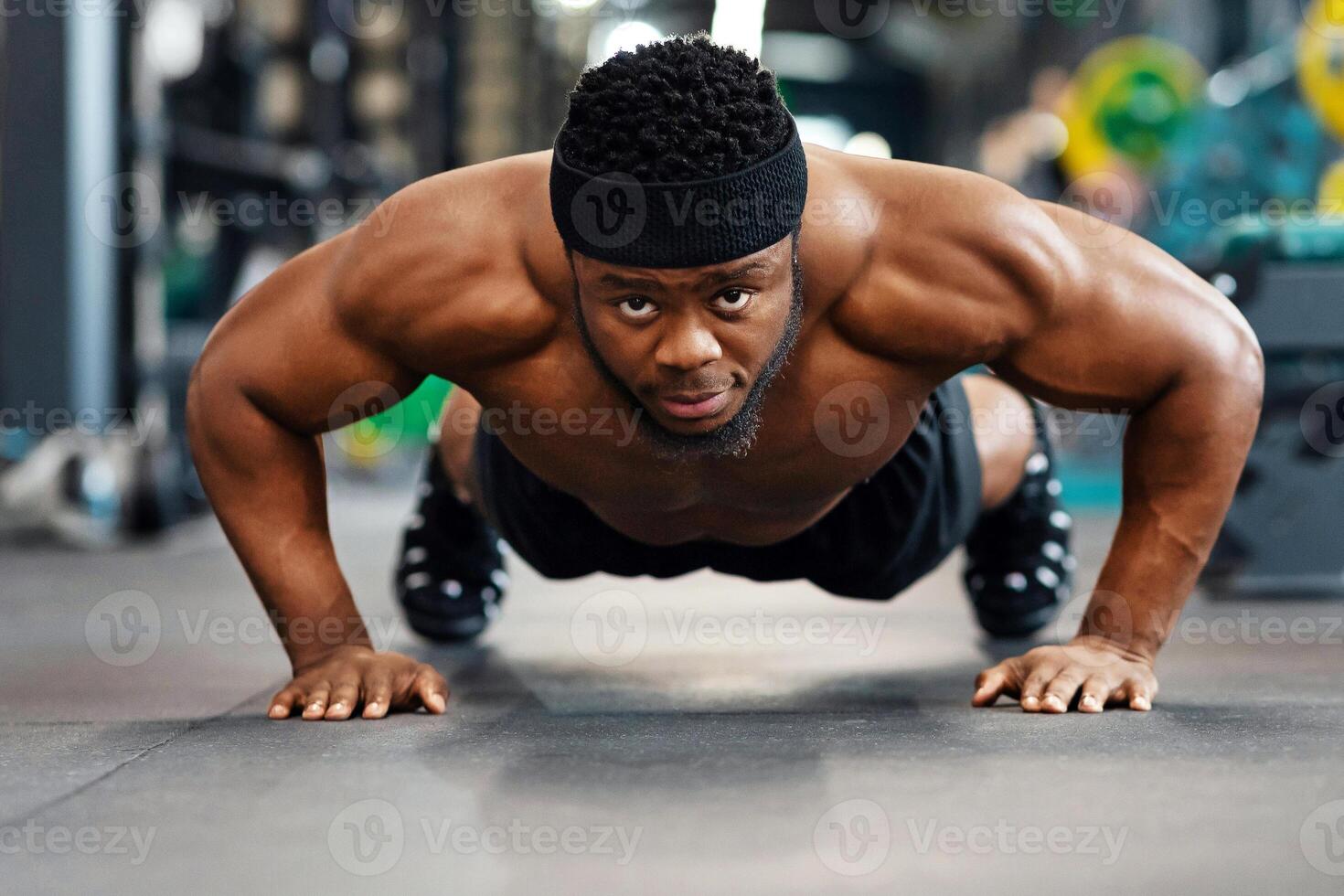Push-ups, press-ups workout. Muscular african american shirtless guy athlete building up muscles, black man training on floor at modern gym, doing push-ups and looking at camera photo